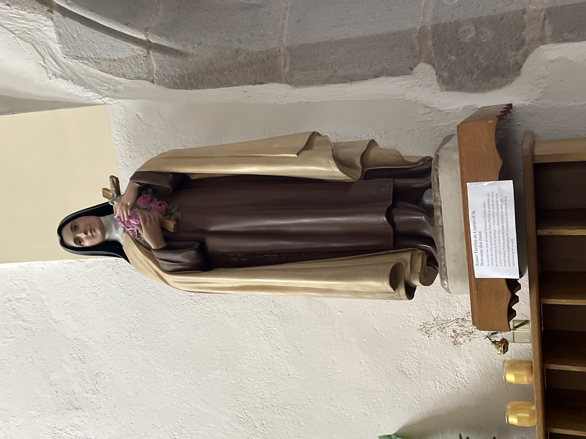 The image shows a statue of Saint Thérèse of Lisieux in a likely indoor location, probably a church or religious setting in Chambœuf, France. The statue, with dark hair and a tan cloak, holds a bouquet of pink roses and a cross. A printed card next to it provides information about Saint Thérèse and the "Novena of Roses" in French. Below the statue is a small wooden shelf with dried flowers and votive candles. The statue is placed against a white wall with architectural details, possibly a stone structure.