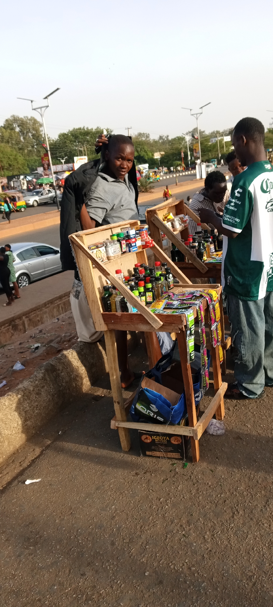 The image depicts a bustling street scene in Jos, Nigeria, focusing on several individuals engaged in street vending and shopping. In the foreground, two wooden stalls are set up by the roadside, filled with numerous small bottles, likely containing traditional medicines, bitters, or perfumes, along with packets of snacks or sweets, some labeled "KRUSH," hanging from the sides.
A man in a grey polo shirt and black jacket stands behind the left stall, looking towards the camera with a neutral expression. Another man, wearing a green and white jersey with text "Omnibus de México" and "SANATORIO ESPAÑOL," stands to the right, facing the stalls, seemingly browsing or purchasing items. A third man in a checkered shirt is visible behind the right stall, also looking at the merchandise.
The background reveals a busy street with cars and a motorized tricycle (keke napep) in motion. Trees, utility poles, and streetlights line the road, suggesting an urban or semi-urban environment. The lighting indicates it is daytime, possibly late afternoon, with clear skies. Underneath the stalls, a blue bag with "ORIS" visible and a cardboard box labeled "IGBOYA BITTERS" are present, further specifying the types of products being sold. The scene captures a typical moment of daily commerce and interaction on a Nigerian street.