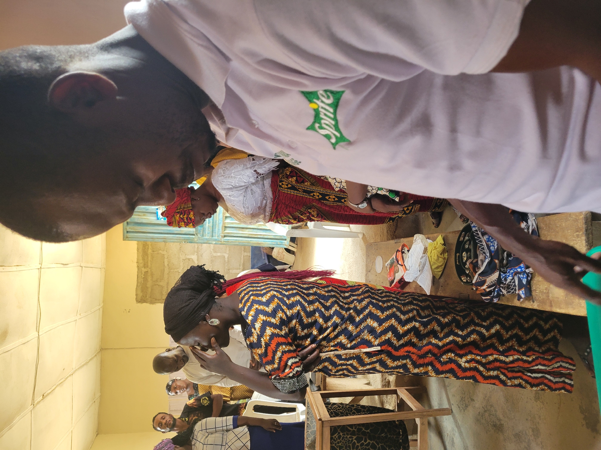 The image shows a gathering of people indoors, potentially in Madalla, Nigeria, though this cannot be forensically confirmed. People are wearing various types of clothing with different patterns, including a shirt with a "Sprite" logo. One woman is wearing a patterned dress and a head covering, holding her face with her hand. Other individuals are seated and standing. The environment appears to be a simple room with painted walls. There are chairs and other objects scattered around.