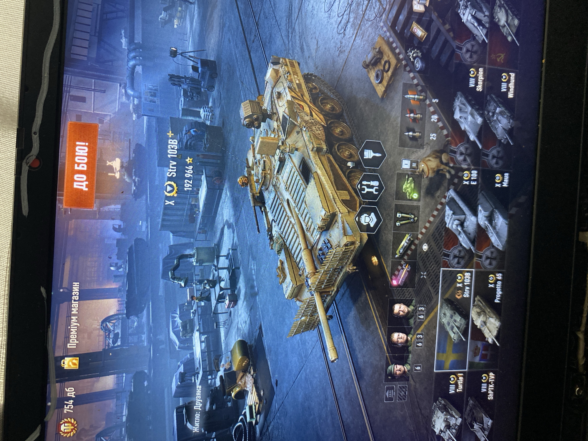 The image shows a screenshot of a video game, likely a tank simulation or combat game, displayed on a laptop screen. The game interface features a hangar or garage scene with several tanks visible, including a prominent golden tank in the foreground. Various UI elements, such as buttons, menus, tank information, and crew portraits, are present. The game displays text in what appears to be a Slavic language. In the top left corner, there is an icon with the number "754 дб" and another icon with "Преміум магазин". A large orange button in the upper center reads "ДО БОЮ!". The environment depicted in the game is an industrial setting with machinery and structures. There are no visible elements in the image that indicate a specific location in the city of Szczecin, Poland, or any real-world setting outside of the game itself.