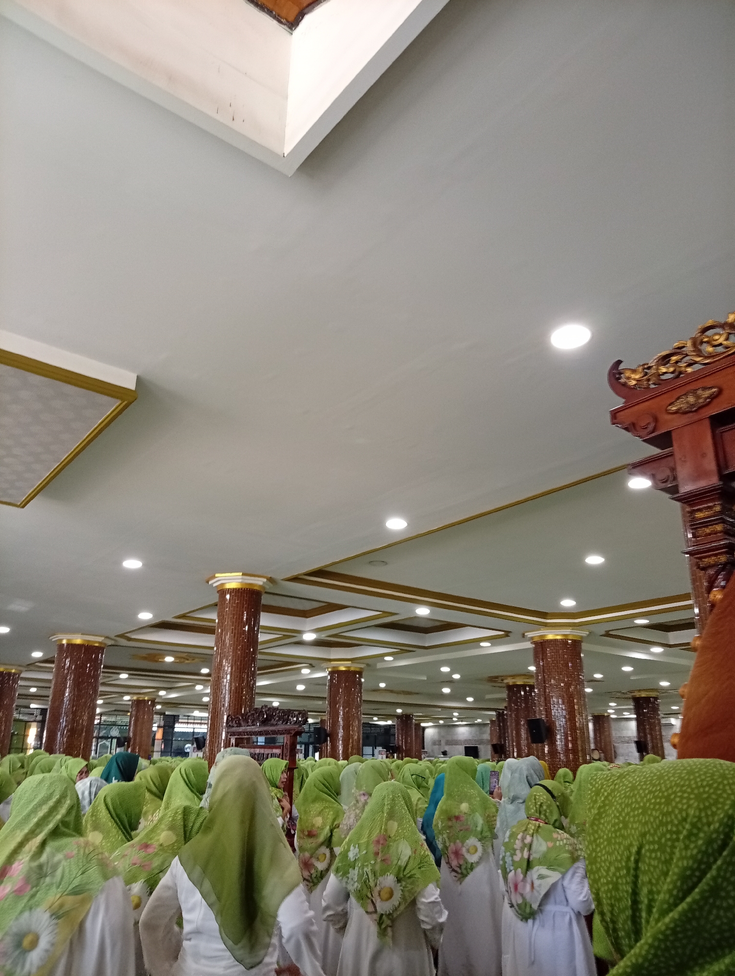 The image depicts a gathering of people, primarily women, inside a large, ornately decorated indoor space. The architecture features high ceilings with recessed lighting and decorative gold trim. Large, cylindrical columns with textured brown surfaces and gold capitals are visible, supporting the ceiling structure. In the foreground and mid-ground, many individuals are wearing light-colored clothing and green headscarves, some with floral patterns. They appear to be facing forward, suggesting they are attending an event or service. The overall atmosphere is one of a communal gathering, possibly in a religious or cultural setting. This scene is located in the city of Sidoarjo, Indonesia, a region known for its diverse cultural and religious practices, and this venue likely serves as a significant location for such events within the community.