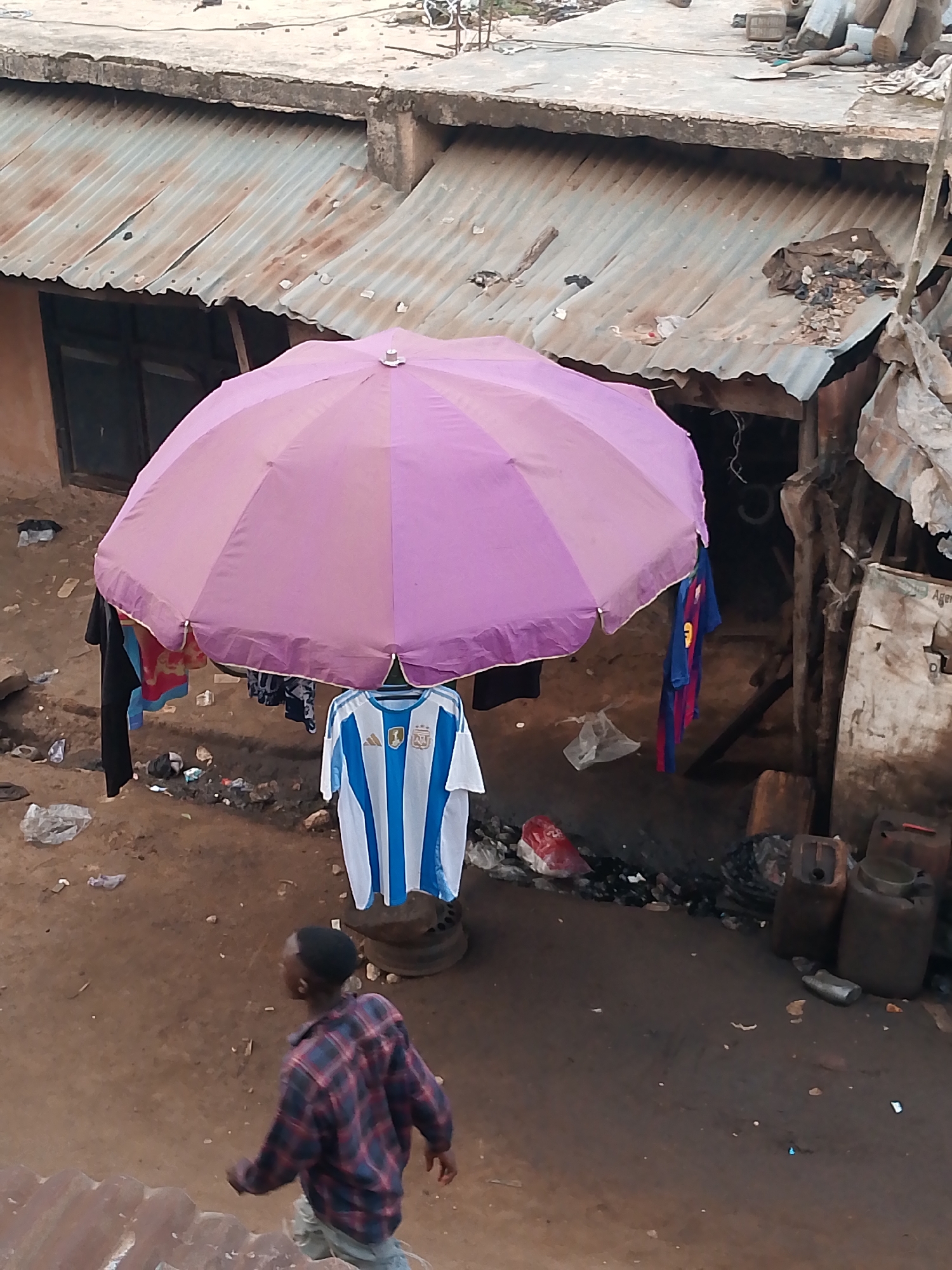 The image shows a scene in Bukuru, Nigeria. An individual is walking away from the viewer. Underneath a large, purple umbrella, clothes, including a blue-and-white striped soccer jersey, hang on hangers. The background shows a dilapidated building with a corrugated metal roof. Debris and trash are scattered on the ground around the building and under the umbrella. Several dark containers stand near the right edge of the frame. The light suggests an overcast day.