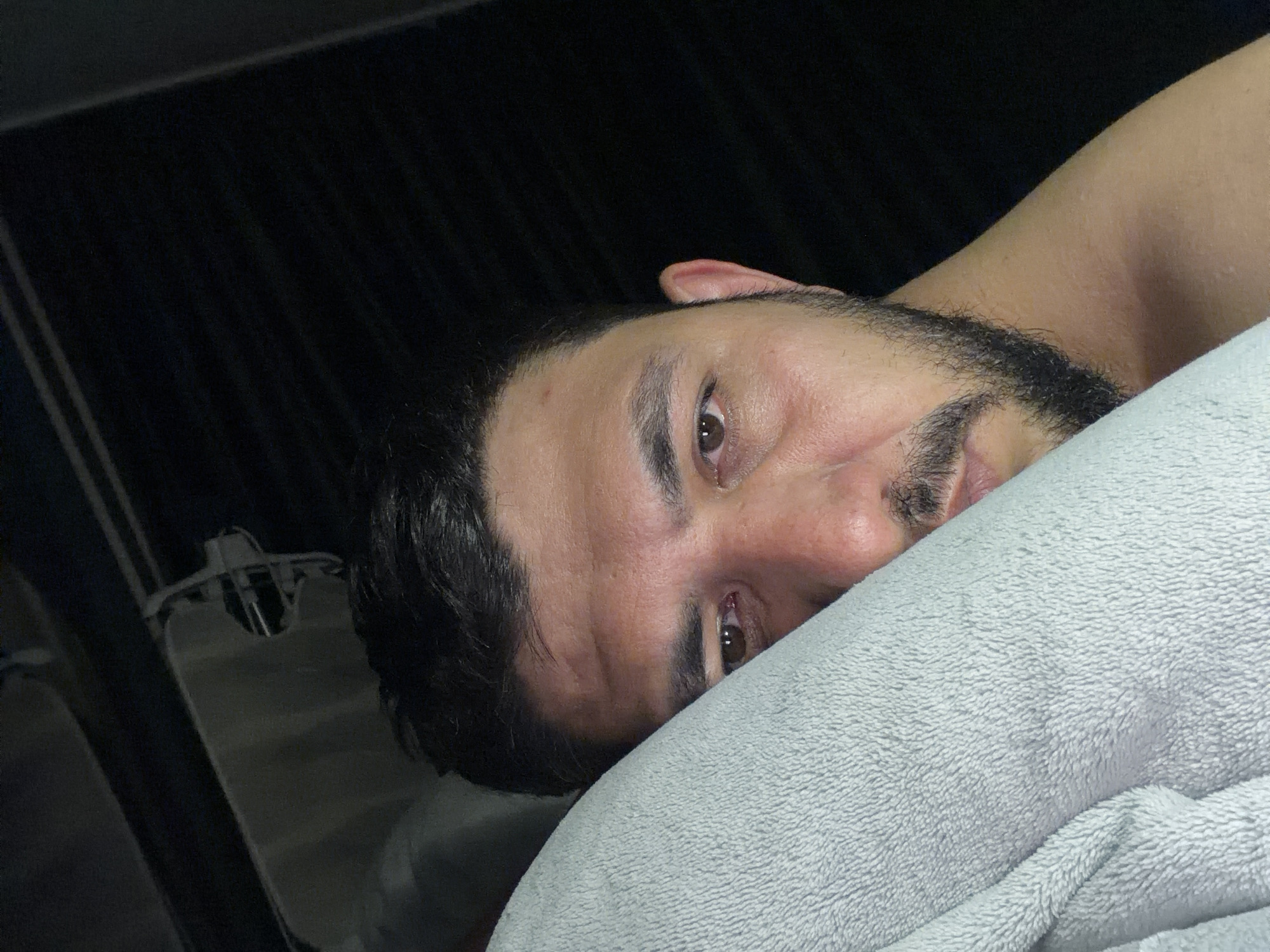 A close-up, angled shot depicts a person lying in bed. The individual has dark hair and a beard, and their face is partially obscured by a light gray blanket or pillow. The person is facing the camera, with one eye more visible than the other. The background is dark, with some indistinct shapes suggesting furniture or clothing. The location is not discernible and there is no evidence indicating this image was taken in Krasnoyarsk, Russia.