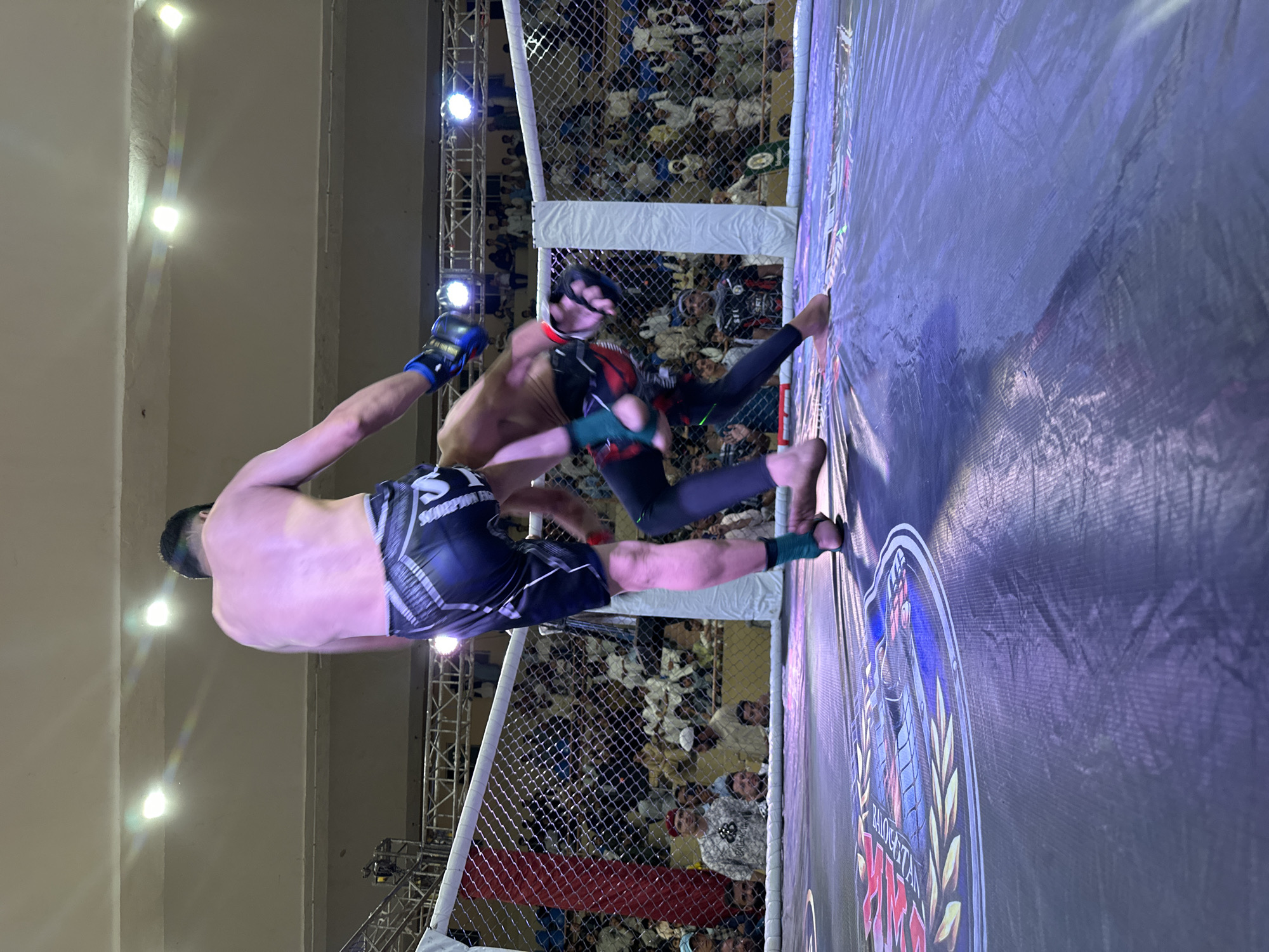 The image depicts a mixed martial arts (MMA) fight taking place inside a caged octagon. Two male fighters are engaged in combat. One fighter, wearing black shorts and blue gloves, appears to be in mid-air. The other fighter is wearing dark pants and headgear. The fight is taking place in front of a large crowd. The octagon is situated in a well-lit area. A banner with the words "BALOCHISTAN MMA" is visible. Based on the logo present, the location is Quetta City Tehsil, Pakistan.