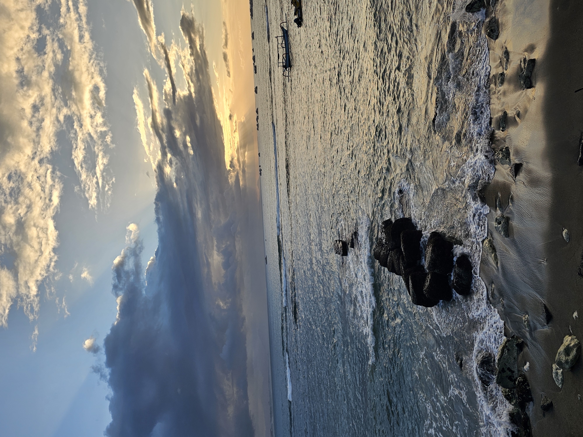 This image depicts a scenic coastal view at sunset in Denpasar, Indonesia. The sky is a dramatic blend of deep blues and grays with shafts of golden sunlight breaking through the clouds. The sea is a tranquil blue, with gentle waves lapping at a dark, rocky shoreline. In the distance, several small boats are visible on the water, and a larger boat with an outrigger is closer to shore. The beach itself is dark sand, scattered with rocks and pebbles. The overall atmosphere is peaceful and serene, capturing the beauty of a tropical sunset.