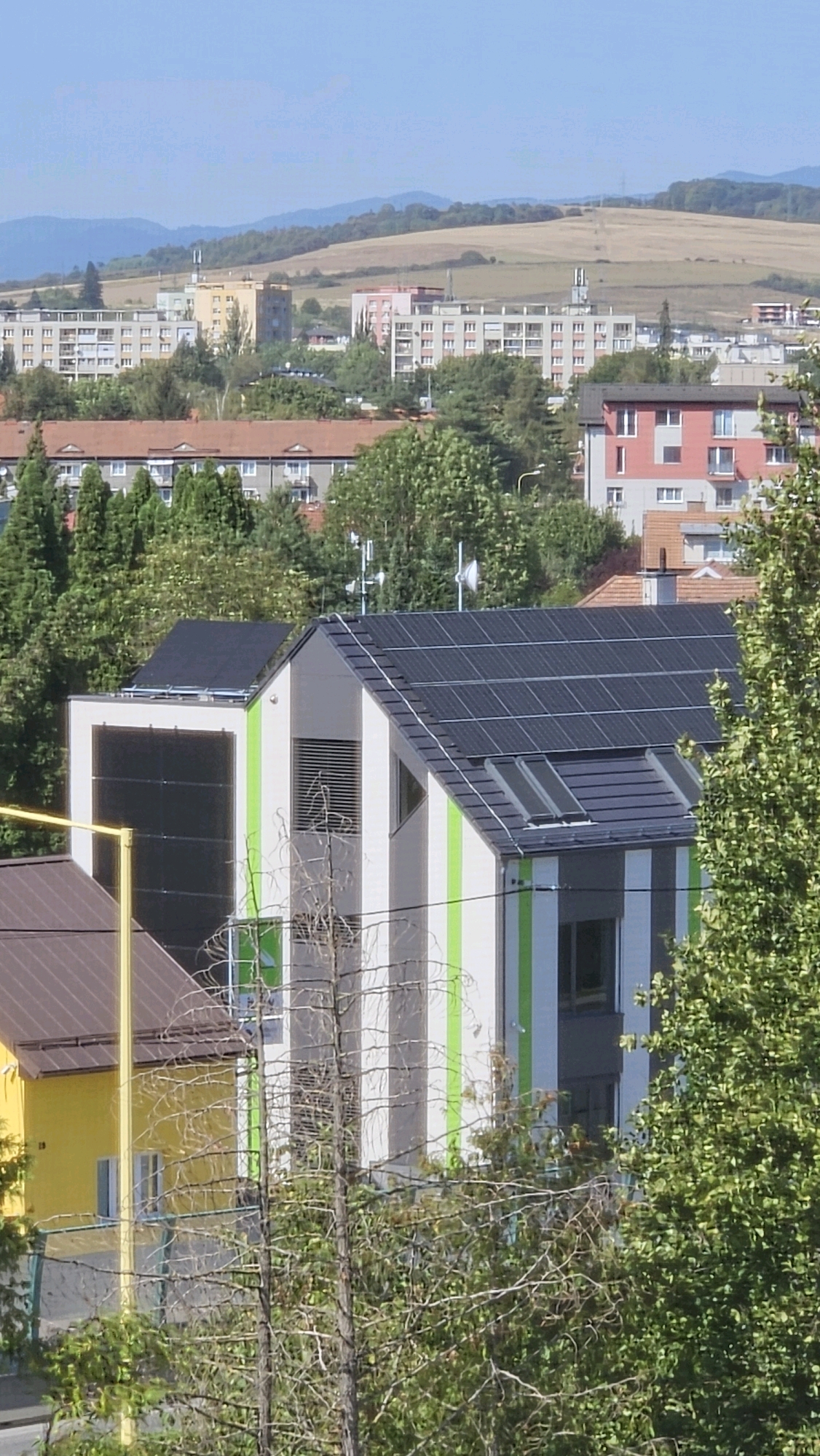 The image depicts a view of residential buildings and surrounding greenery in Prešov, Slovakia. In the foreground, a modern building with solar panels on its roof is visible, featuring a white facade with green vertical stripes. Adjacent to this is a yellow building with a brown roof. Behind these, a collection of taller apartment buildings, typical of socialist-era architecture, form the middle ground, interspersed with trees. Further in the distance, rolling hills with fields and some scattered buildings complete the urban landscape under a clear blue sky. There are no people or animals visible in the scene, and no specific actions or interactions are occurring. The overall impression is one of a suburban or residential area within the city.