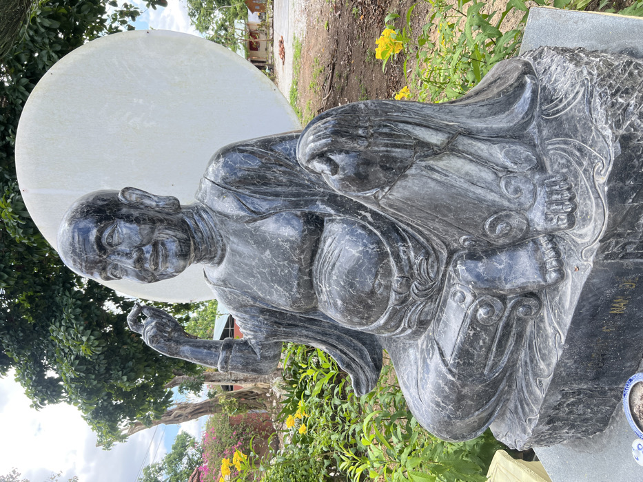 A dark grey, mottled stone statue depicting a bald, elderly male figure with a prominent belly is centrally positioned outdoors in Đăk Mar, Vietnam. The figure is seated with crossed legs, wearing a draped robe, and has a smiling expression with large earlobes. His right hand is raised, holding a small, spherical object between the thumb and forefinger, while his left hand rests on his left knee. The statue stands on a dark stone base with "NAN D" inscribed in gold. Directly behind the statue is a large, weathered white circular disc. The immediate environment includes green foliage, yellow flowering shrubs, and distant pink flowers. In the background to the right, small buildings with red roofs are visible near a dirt or gravel path. The sky is partially cloudy.