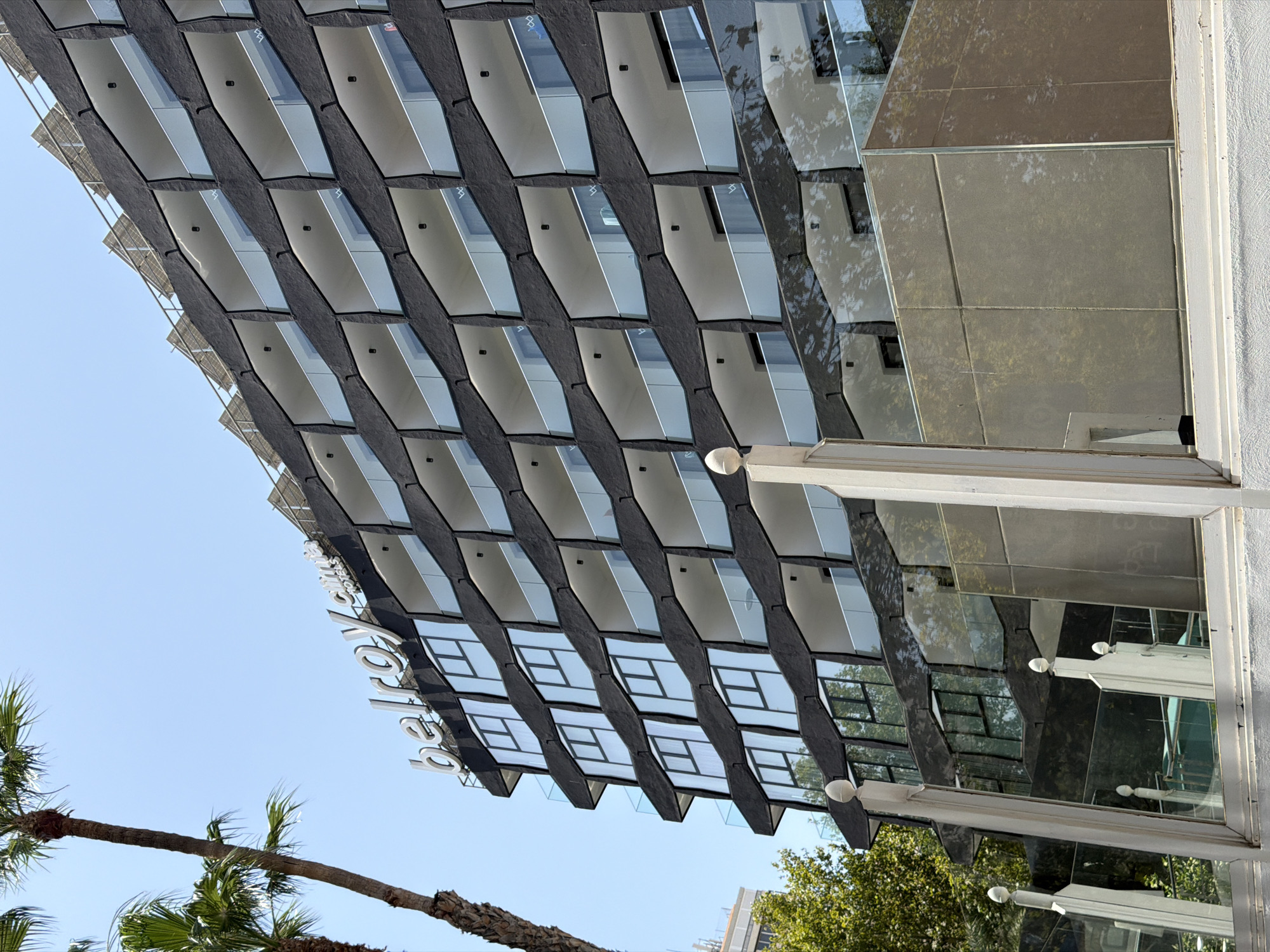 The image shows the exterior of the Belroy hotel, a resort located in the city of Benidorm, Spain. The hotel features a modern architectural design with a dark, textured facade and multiple levels of balconies with glass railings. Solar panels are visible on the rooftop. Palm trees frame the left side of the building, and a bright, clear sky serves as the backdrop. In the foreground, there is a glass barrier reflecting the hotel's facade and surrounding greenery. The scene captures a sunny day in a tourist destination.