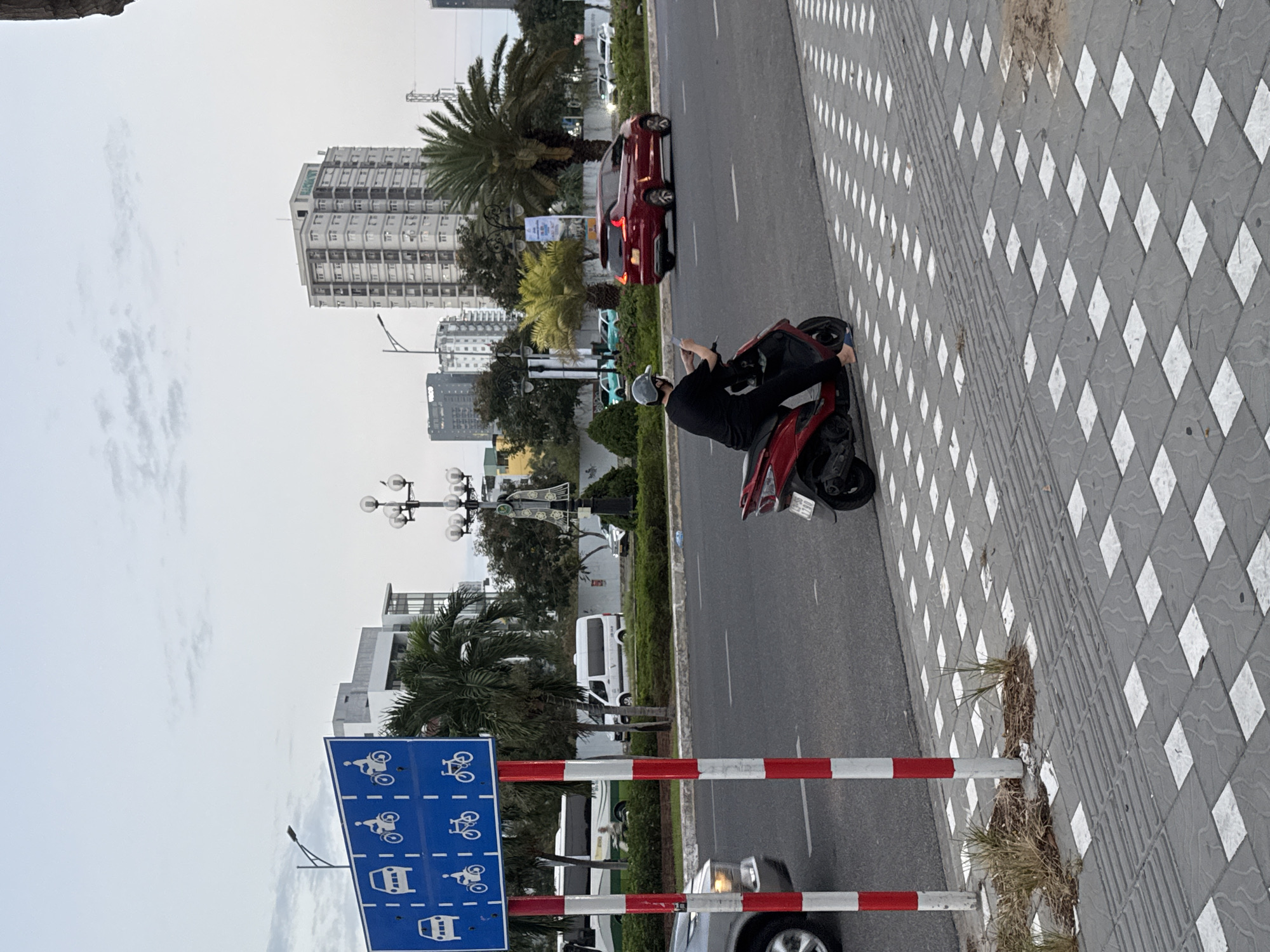 In Đà Nẵng, Vietnam, a street scene unfolds with a traffic lane and a pedestrian walkway. A blue sign with white pictograms indicates traffic regulations. The sign displays symbols for cars, motorcycles, and bicycles arranged in a grid. Two red and white striped poles support the sign. The street is paved and a red scooter with a rider wearing a black shirt and a grey cap is visible on the right. The rider is holding up a mobile phone, possibly filming or taking a picture. A red car is seen driving on the road in the distance. The pedestrian area is paved with interlocking tiles, some of which are white squares. In the background, buildings and palm trees are present, suggesting an urban environment. The sky is overcast.