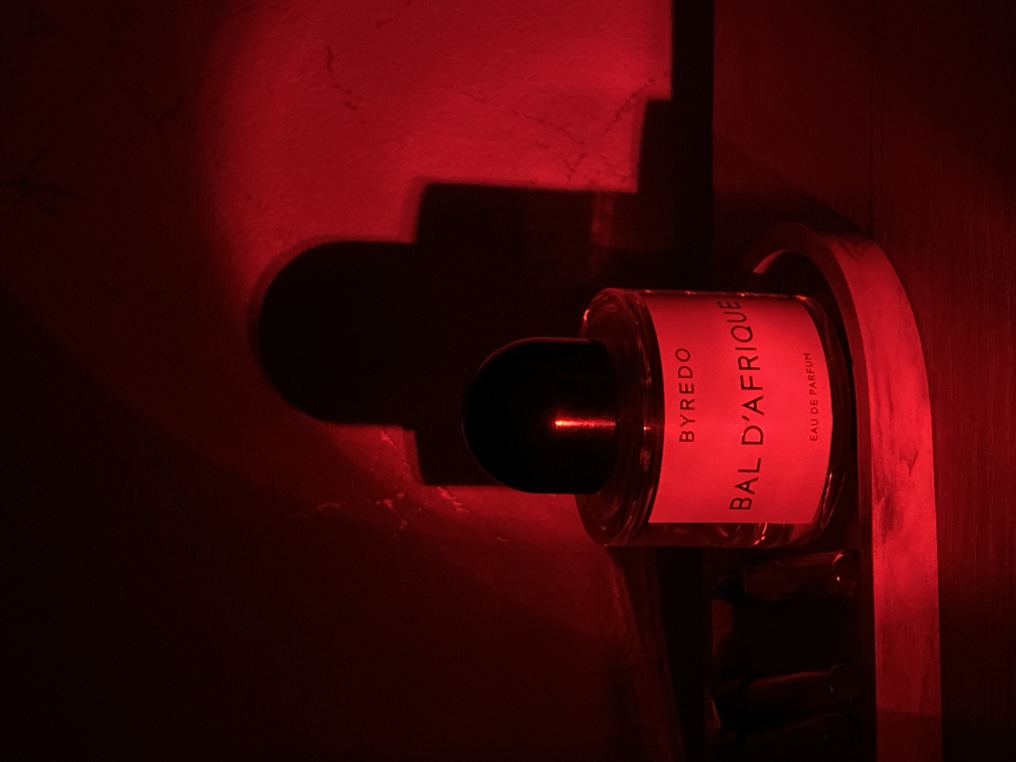 The image displays a Byredo "Bal d'Afrique" Eau de Parfum bottle, oriented horizontally, against a deeply red-lit background. The setting appears to be indoors, possibly on a wooden shelf or within a niche, with a textured wall behind it. The bottle itself is clear glass with a prominent red label featuring white uppercase text that reads "BYREDO" at the top, "BAL D'AFRIQUE" in the center, and "EAU DE PARFUM" below. It has a distinctive black, cylindrical cap.
The scene is dominated by strong red artificial lighting, which creates a dramatic, moody atmosphere. This light source casts sharp, angular shadows of an architectural element, possibly a decorative wall feature, onto the background wall. The shadows appear geometric, suggesting steps or tiered shapes. A thin, horizontal streak of red light is visible reflecting off the black cap of the perfume bottle. The lighting highlights textures on the wall and the wooden surface where the bottle rests. No people are present, and no specific activity is occurring. The overall impression is one of stylized product photography, emphasizing the luxury item under unique lighting conditions in Sochi, Russia.