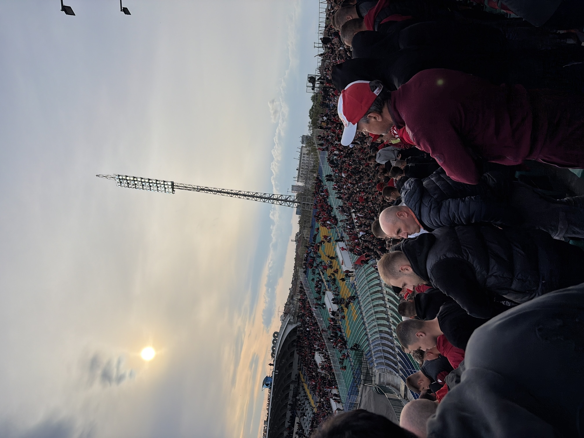 The image captures a crowded outdoor stadium scene at sunset, likely during a sporting event. Numerous spectators, primarily men, are visible from behind and the side, filling the tiered stands. In the mid-ground, a man in a maroon jacket and a red and white baseball cap, which has the word "VICTOR" visible, is prominent. Other individuals in the foreground are seen wearing dark, often padded, jackets.
The stadium features multicolored seating sections in green, yellow, and blue. A tall, industrial floodlight tower, equipped with multiple lights, stands prominently against the sky on the left side of the frame, with other smaller floodlights visible higher up. The low-lying sun casts a bright, warm glow through scattered clouds, indicating late afternoon or early evening. The sky is partly cloudy but luminous around the sun. The collective gathering of the audience suggests an event such as a football match. The scene is set in Sofia, Bulgaria.