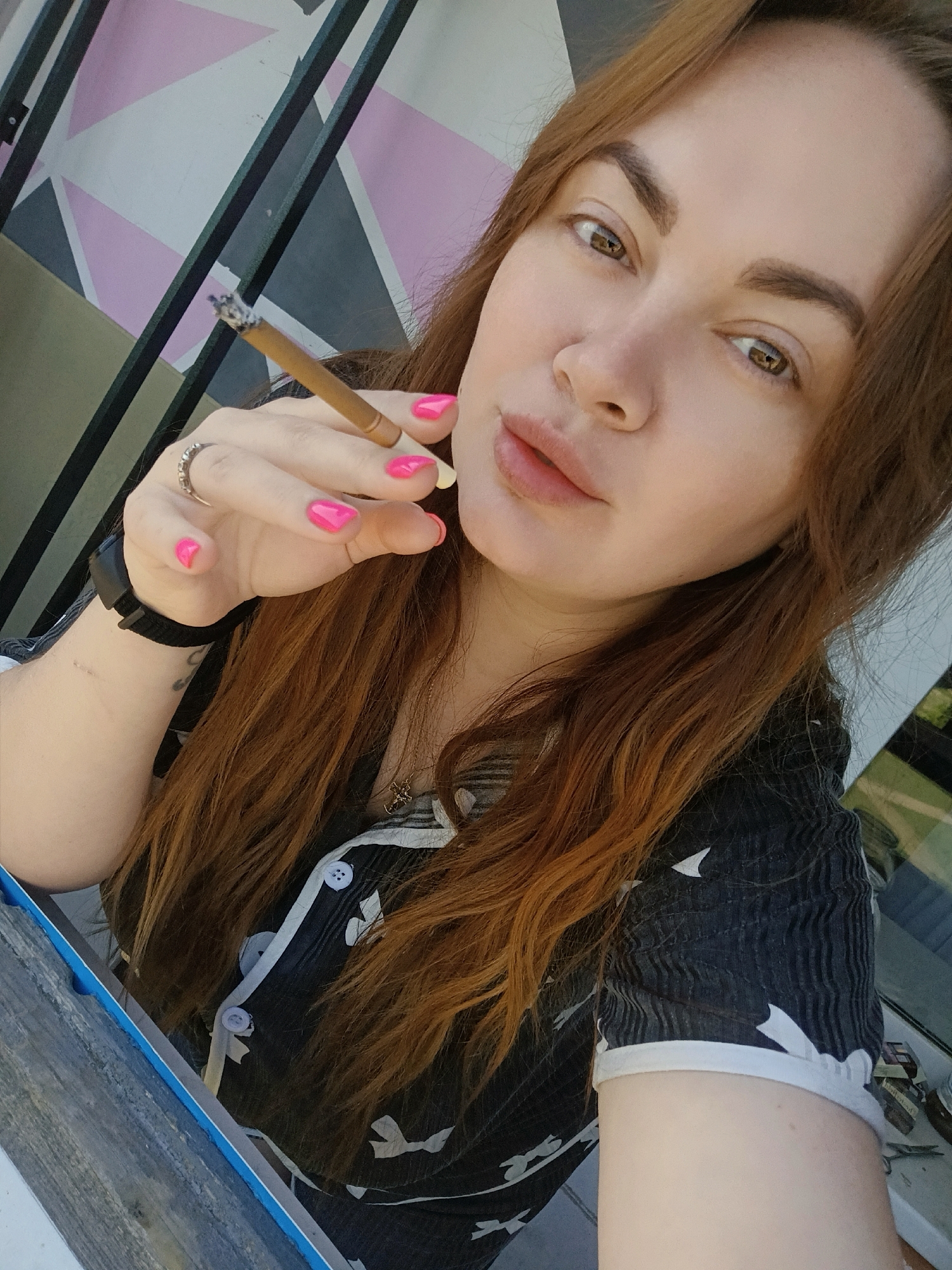 A light-skinned woman with brown hair and brown eyes is holding a lit cigarette in her right hand. She is wearing a black and white patterned top with white buttons. The background includes a wall with geometric shapes in pink, white, grey, and black, and a dark-colored metal railing. She has a black watch on her left wrist and a silver ring on her right ring finger. The photo was taken in Chaikovsky, Russia.