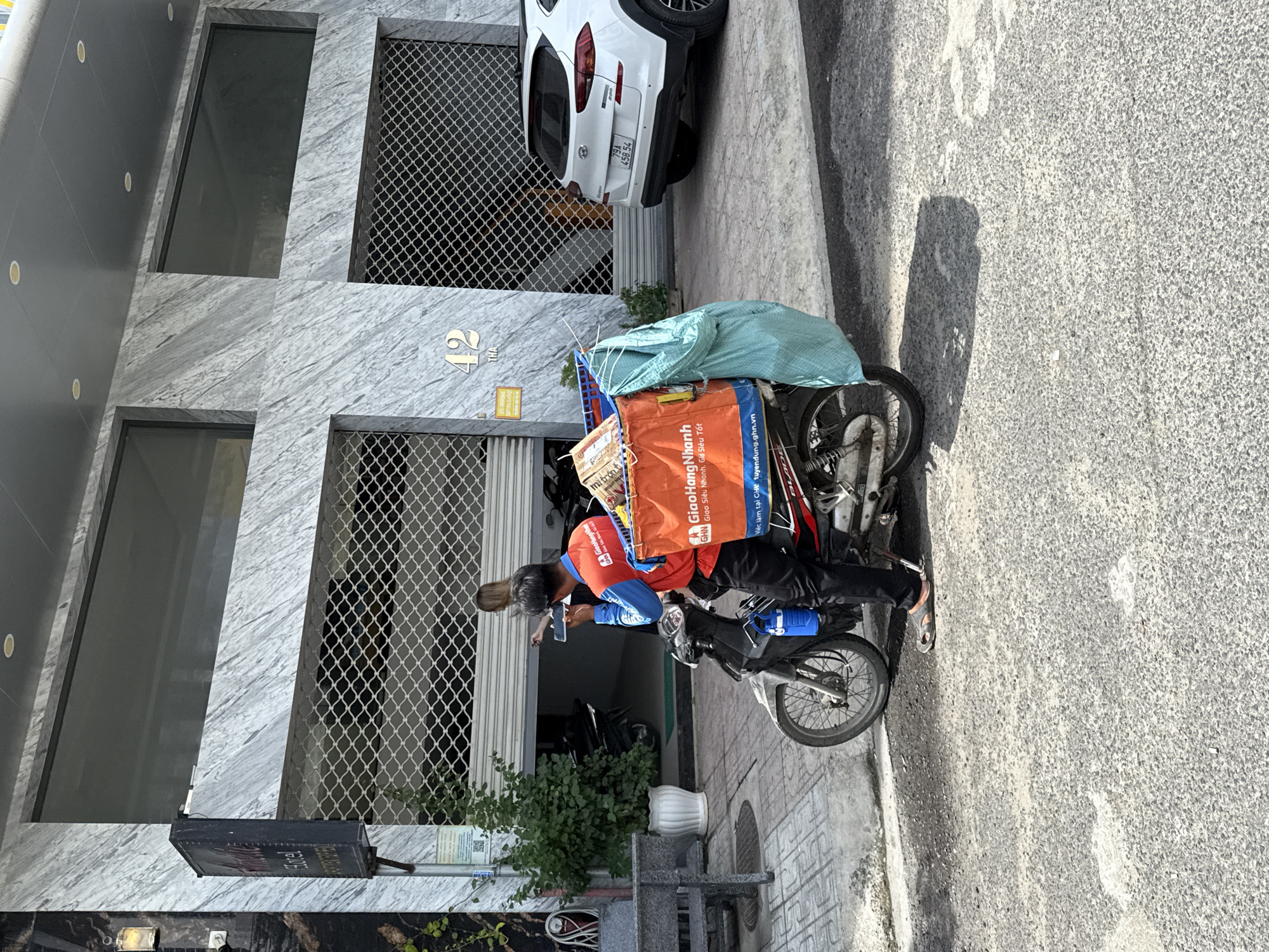 An outdoor scene in Bac Nha Trang Ward, Vietnam, shows a man dressed in an orange vest and black pants standing next to a motorcycle. The man is holding a mobile phone to his ear and appears to be speaking. The motorcycle is equipped with a large orange bag attached to the rear, containing several boxes and a light blue tarp. The building in the background features a modern facade with gray marble-like cladding and large metal roll-up doors. A white SUV is parked on the right side of the frame. The ground is paved with textured concrete.
