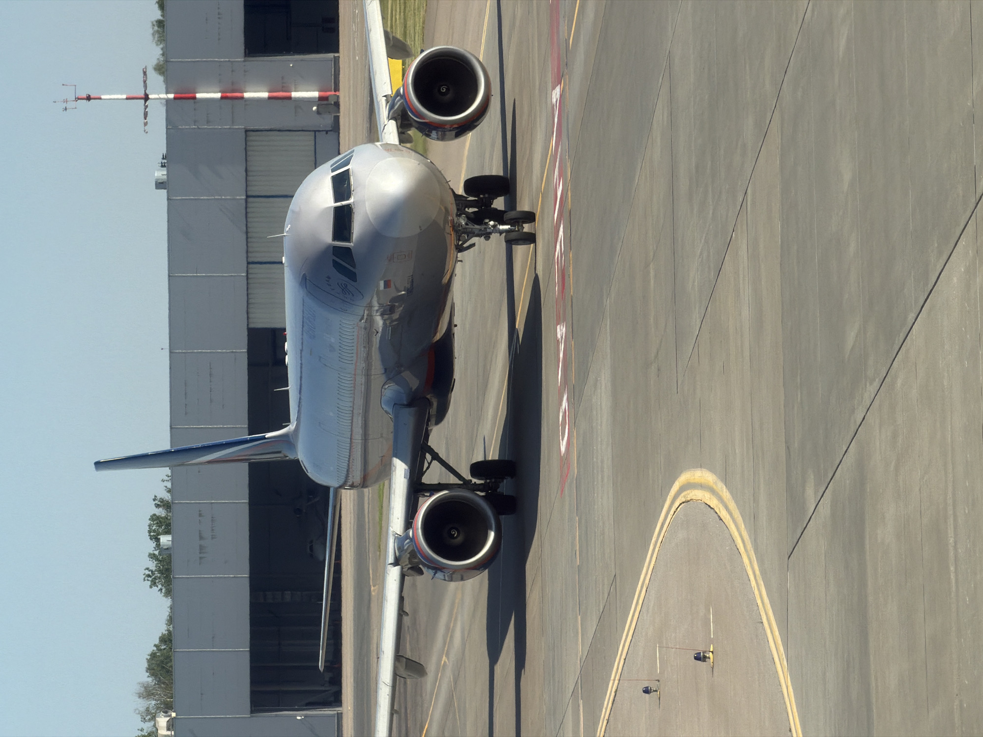 An Airbus A320 aircraft, bearing Russian flag insignia and the Aeroflot livery, is positioned on an airport tarmac. The aircraft, a twin-engine jet with its nose angled slightly to the right, is illuminated by daylight. Its landing gear is visible, with the front wheels turned. In the background, a large hangar-like structure stands, its entrance open, revealing another, smaller aircraft inside. To the right of the hangar, a tall, red and white striped pole, likely a meteorological instrument or marker, is present. The tarmac is marked with yellow and red lines, indicating taxiways and runways. The environment appears to be an airport, possibly Sheremetyevo International Airport, located in Khimki, Russia. The sky above is clear and blue.