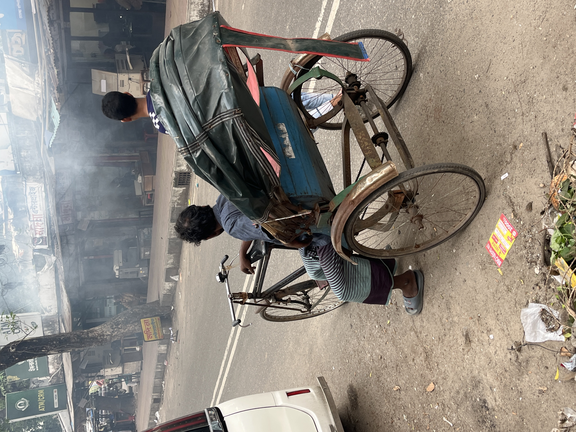 The image, taken in Dhaka, Bangladesh, depicts a rickshaw puller resting. He is sitting on the seat of his rickshaw, which has a dark green, partially covered canopy. He wears a striped shirt and dark shorts with a turquoise stripe. A passenger, wearing a blue and white checkered shirt, sits behind him under the canopy. The rickshaw, displaying signs of rust and wear, is parked near a curb on a street with a white dividing line.
To the right of the rickshaw, there is trash and a flyer on the ground. In the background, across the street, are buildings with signs in Bengali, including ATM and banking logos. Smoke or steam obscures the view of the buildings. The front of a white vehicle is visible at the bottom of the image, partially obscuring the bottom of the rickshaw puller’s bike and wheel.