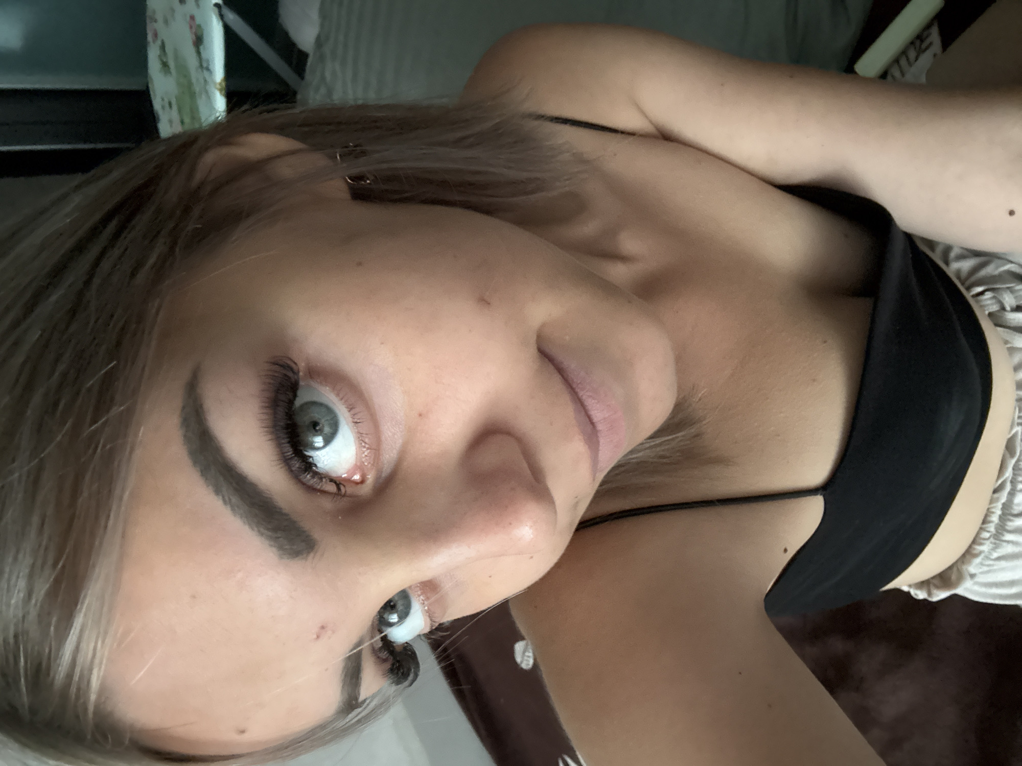 A young woman with light brown hair and grey eyes is captured in a close-up, selfie-style shot. She is wearing a black camisole and light-colored shorts. Her makeup includes prominent false eyelashes and defined eyebrows. The background is blurred, showing what appears to be a domestic setting with bedding and possibly an ironing board. The image is taken indoors, with soft lighting. There is no indication of the city of Yuzhno-Sakhalinsk, Russia.