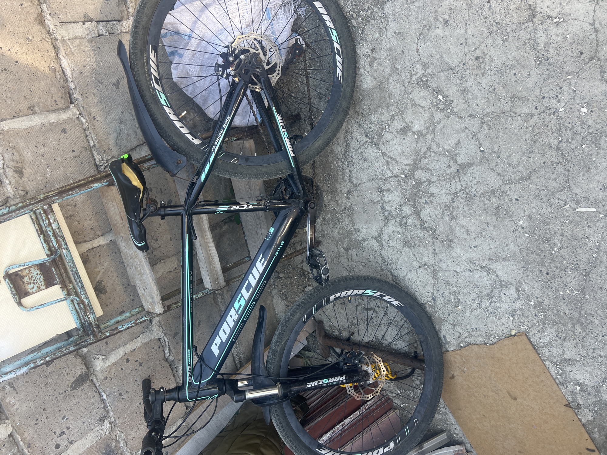 An outdoor, eye-level, full shot shows a black and teal mountain bike leaning against a concrete wall. The bicycle has "PORSCHE" written in white and teal on the frame and wheels. The front of the bike is on the left, with its wheel turned slightly to the right. The handlebars are black with black grips. The bike has black fenders and disc brakes. The rear wheel is on the right, also with "PORSCHE" visible on the tire. A worn, black seat with yellow padding is visible. The bike rests on a cracked, gray concrete surface. Behind the bike is a rough concrete wall with some weathered wooden planks and a rusty, blue metal structure. The scene is illuminated by natural sunlight.