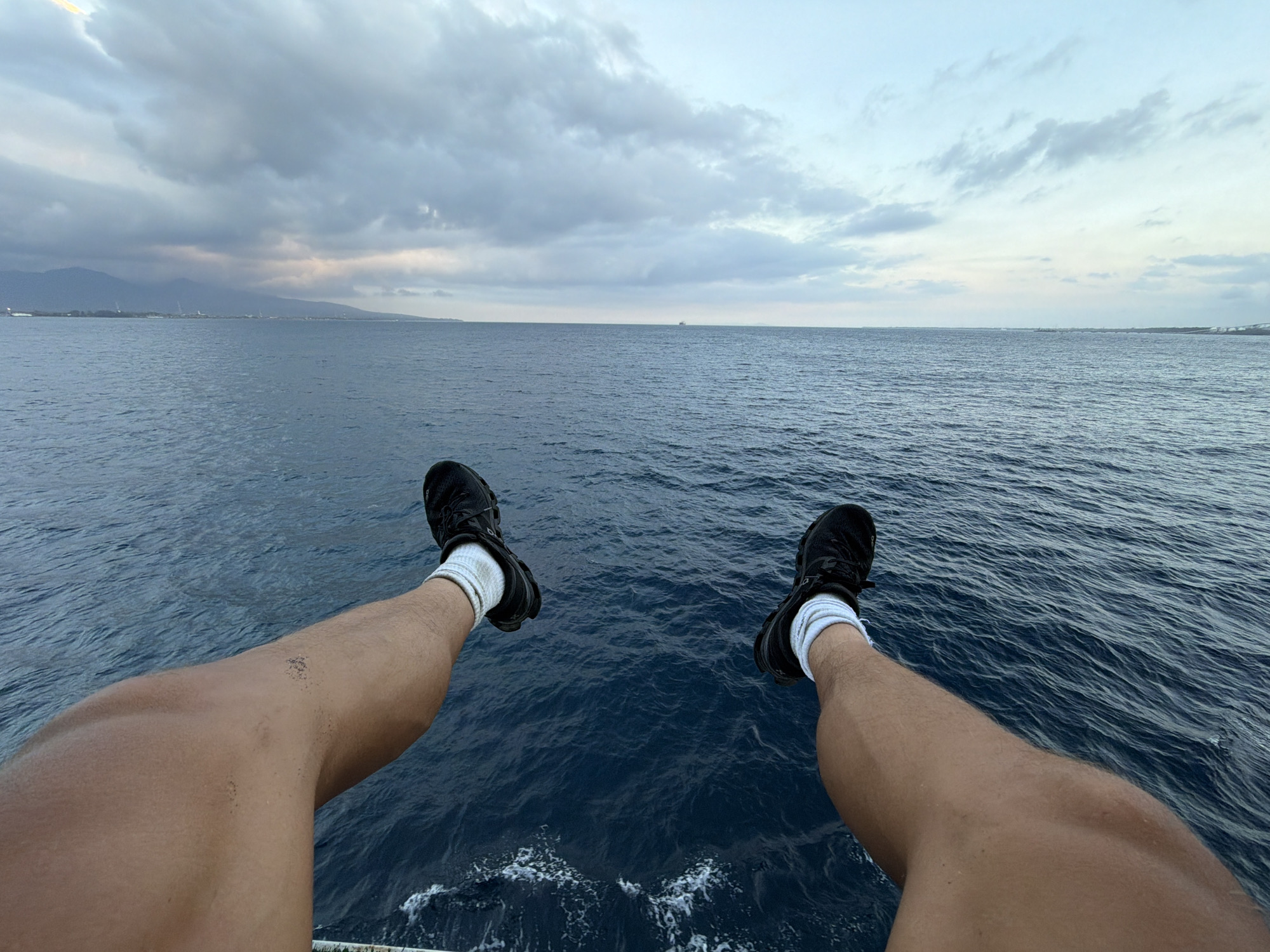 A first-person perspective shot shows a pair of legs and feet dangling over the edge of a boat or structure, with the deep blue ocean stretching out to the horizon. The legs are tanned and bare, with black athletic shoes and white socks visible. The sea is calm, with small ripples and white foam visible where waves are breaking. In the distance, the coastline of Melaya, Indonesia, can be seen, with mountains and a hazy sky. The sky is overcast, with dramatic clouds in shades of gray and hints of pink and yellow as the sun sets or rises. The overall scene conveys a sense of freedom and tranquility.