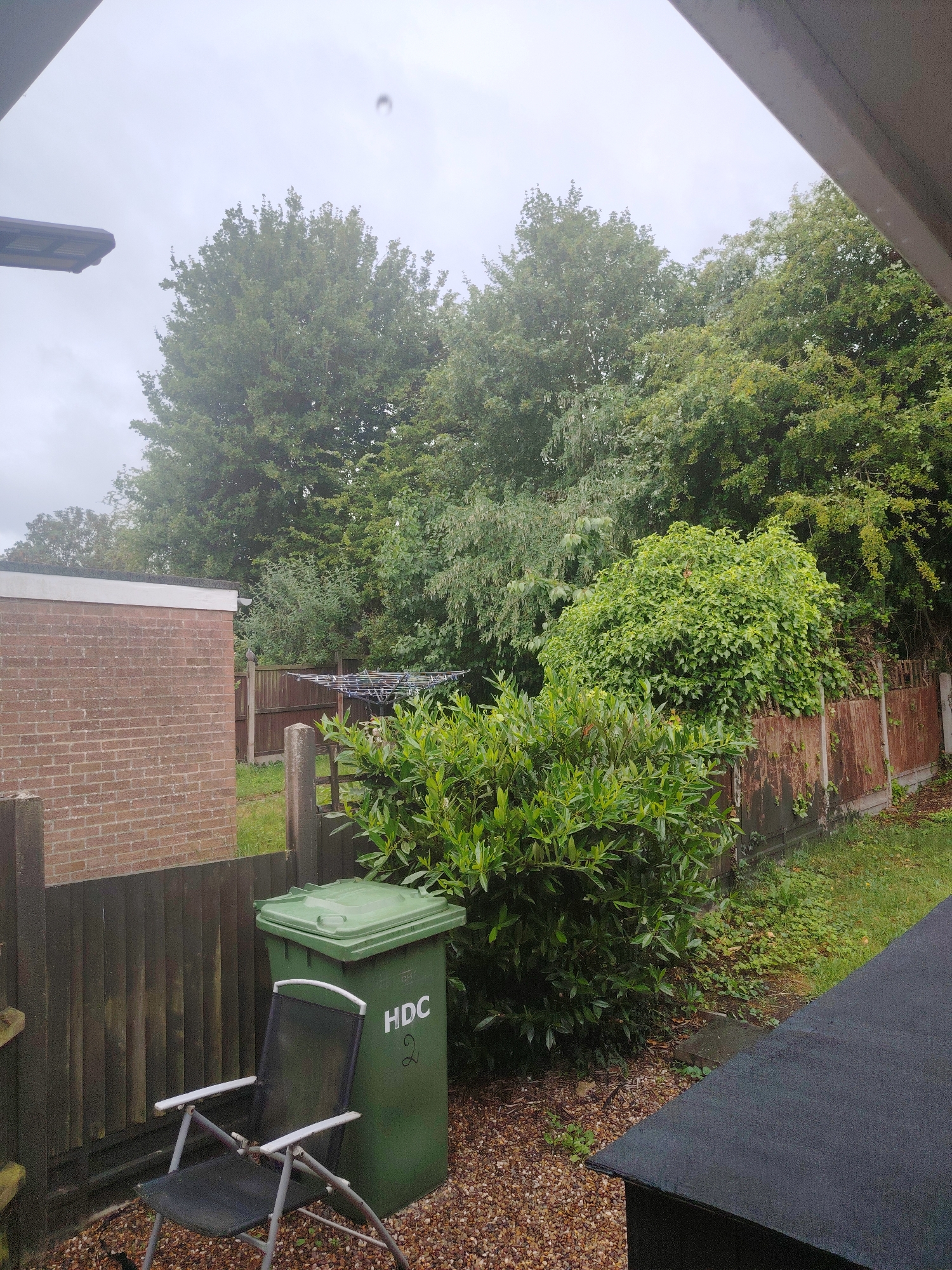 The image depicts a rear garden in Huntingdonshire, United Kingdom, on a cloudy day. In the foreground, a dark grey folding chair with a silver frame sits on a gravel surface. To the right of the chair is a large, green wheelie bin with the letters "HDC" and the number "2" written on its side in white. Behind the bin and chair, a wooden fence extends horizontally across the frame. Beyond the fence, lush green trees and bushes dominate the background. A portion of a brick building with a white roofline is visible on the left side of the image, bordering the fence. A clothes drying rack with clothes on it is positioned further back in the garden, near a weathered wooden fence. The sky is overcast and grey. The image appears to be taken from an elevated position, possibly from an upstairs window or balcony, as indicated by the angled roofline in the top right and left corners.