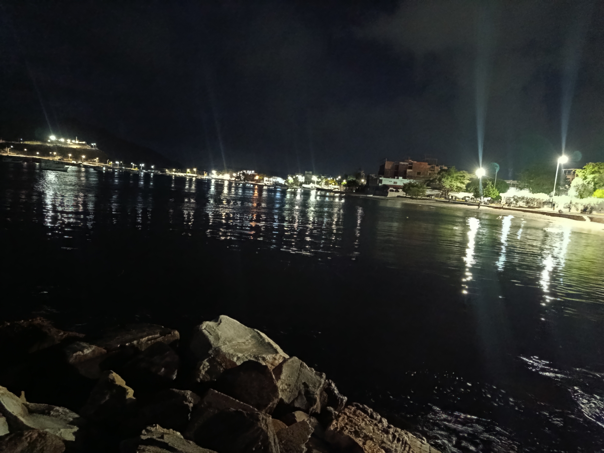 This is a nighttime photograph of the waterfront in Juan Griego, Venezuela. The image is dominated by a dark body of water reflecting the lights from the shoreline. In the foreground, large rocks are visible on the left, leading down to the water's edge. Across the water, the shoreline is illuminated by numerous lights from buildings and streetlamps. A few boats are visible moored near the shore. The sky is dark with some clouds, and the lights from the town cast long reflections on the rippling surface of the water. The overall impression is of a peaceful, illuminated waterfront at night.