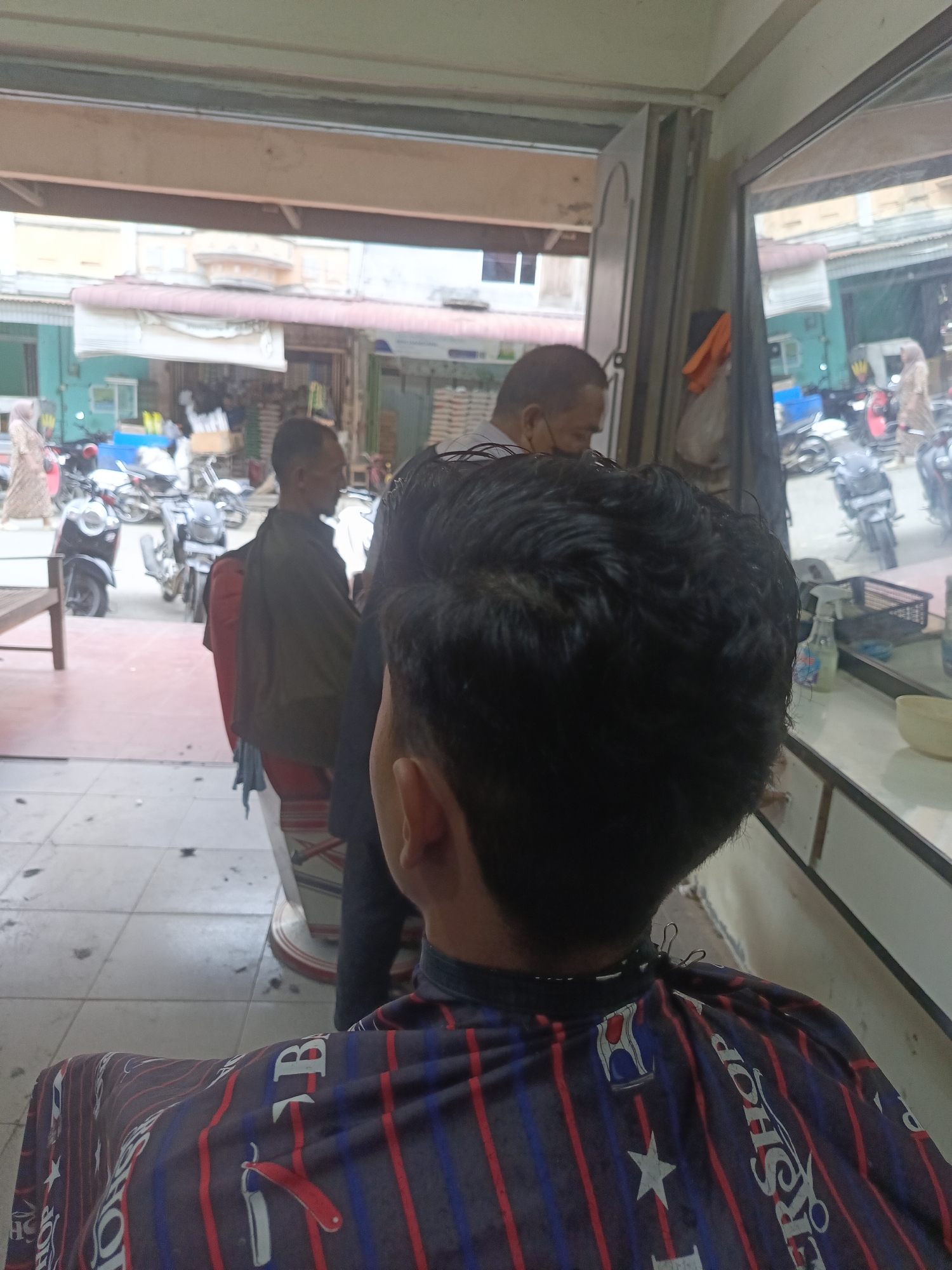 The image depicts the interior of a barbershop located in Lhokseumawe, Indonesia, as evidenced by the "Barbershop" text on the cape. The perspective is from a customer receiving a haircut, looking over their own shoulder at the barber and the surrounding environment. In the foreground, a customer is seated, wearing a cape with a red, white, and blue striped pattern featuring the word "Barbershop" and graphics of scissors and stars. The barber, a man with dark hair and a light-colored shirt, is visible in the background, facing away from the camera and appearing to be in the process of cutting the customer's hair. Another man, also with dark hair and wearing a dark jacket, is seated to the left of the barber.
Through an open doorway and a reflective mirror, a street scene is visible, with several motorcycles parked and people walking. The environment appears to be a bustling urban area. The floor of the barbershop is tiled, with some stray hairs scattered on it, indicating recent activity. A wooden bench is present to the left of the frame. The overall impression is a candid snapshot of a typical barbershop scene in Lhokseumawe.