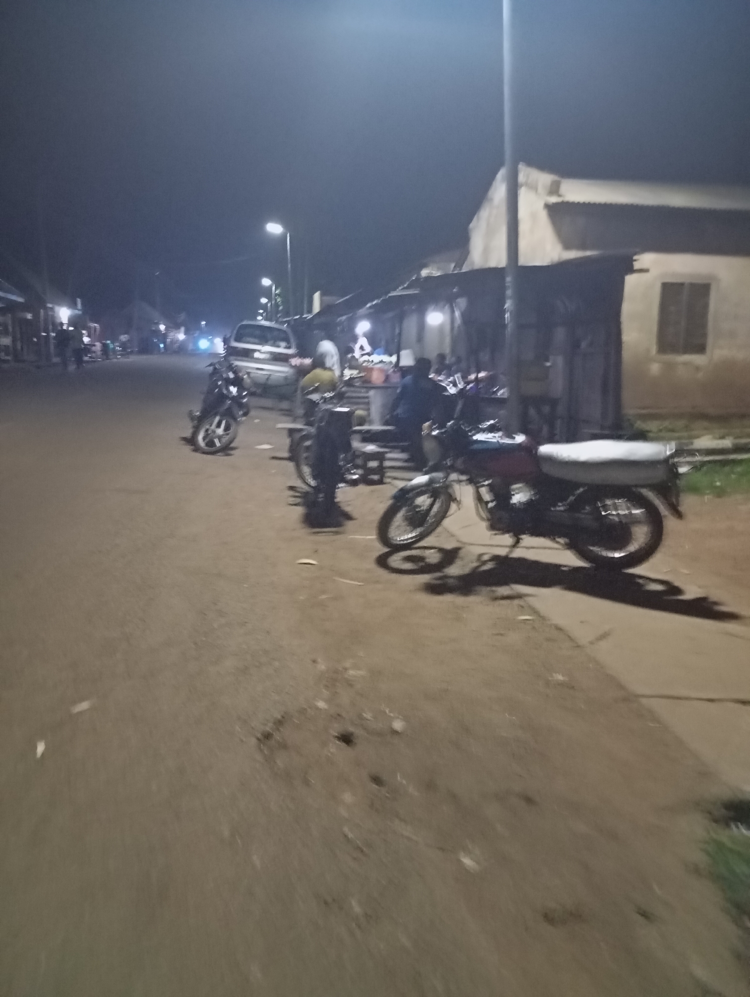 A street scene in Shendam, Nigeria, at night. The street is paved and lined with buildings that appear to be shops or homes. Several motorcycles are parked on the side of the street. In the background, there are lights from buildings and streetlights, as well as vehicle headlights. Some people are visible walking or standing near the shops. The atmosphere is dimly lit, suggesting it is late at night or in an area with limited lighting.