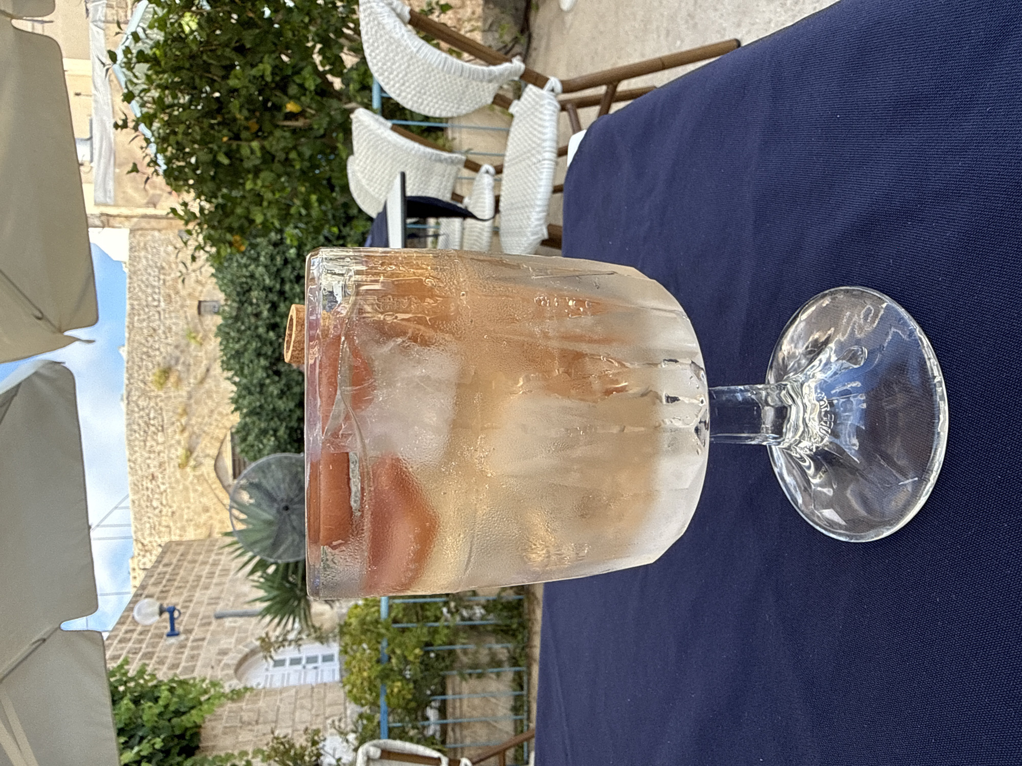 This is a close-up, horizontally oriented, slightly blurry photograph of a clear glass filled with ice and a light-colored beverage, likely a cocktail or infused water, with pieces of orange or peach visible within the drink. The glass has a thick stem and a wide base, and condensation is visible on its exterior. The drink itself appears amber or rosé in color. The glass is placed on a dark blue tablecloth.
In the background, out of focus, are elements of an outdoor setting. There are white woven chairs with curved backs, suggesting a cafe or restaurant patio. Green foliage from trees and bushes is visible, and beyond that, a stone building with a tiled roof and architectural details indicative of the Mediterranean region. A light fixture is mounted on the building. The lighting suggests it is daytime, likely sunny. The overall impression is a relaxed, outdoor dining or lounging experience. No people are visible, and no text is legible in the image.