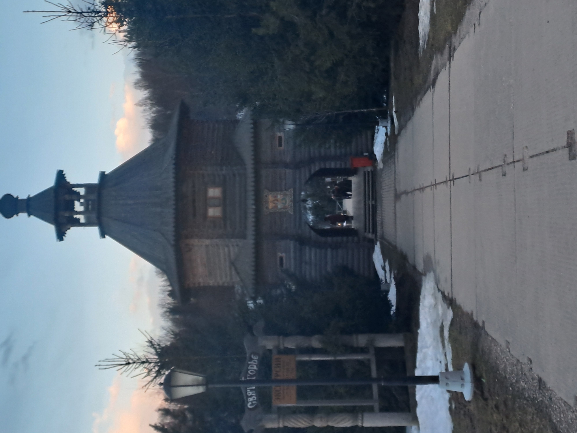 The image shows an exterior view of a wooden building in Vzglyadnevo, Russia. The building features a distinctive onion dome atop a multi-tiered structure. A paved walkway leads to the entrance of the building. Signposts are located near the walkway, but their inscriptions are illegible. Patches of snow are visible on the sides of the walkway and in the background. There are some people visible in the entrance of the building. The sky is visible in the background, displaying hues of blue and light pink. The image is taken from a slightly low angle.