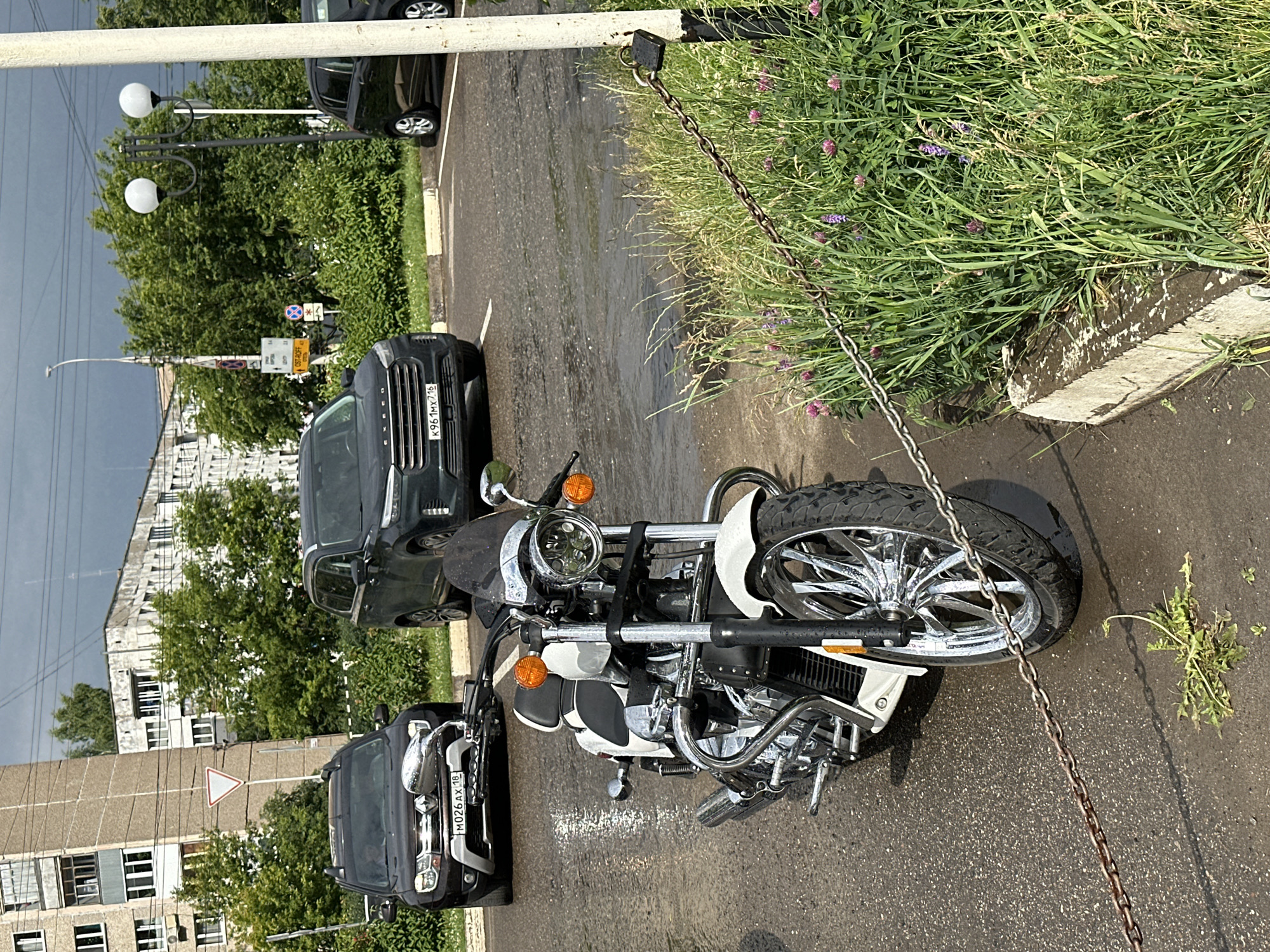 The image captures a street scene in Izhevsk, Russia, on a partly cloudy day. The foreground features a white and chrome motorcycle parked on a wet asphalt surface, possibly due to recent rain, as indicated by puddles and reflections. A rusted metal chain secured by a padlock runs across the lower portion of the image, separating the motorcycle from a patch of overgrown green grass and wildflowers.
In the background, several cars are parked. A dark brown SUV, possibly a Renault Duster with license plate M026AX 18, is visible to the left. Next to it, a dark green SUV, identified by its license plate K961MX 716 as a Chery Exeed, is parked. Further to the right, a dark-colored car, partially obscured, is also present.
The urban environment is further depicted by the presence of apartment buildings and trees. A multi-story apartment building with a light-colored facade and numerous windows stands to the left. Behind the parked cars and vegetation, more trees and structures are visible, suggesting a typical residential or commercial area within the city. A streetlamp with two spherical lights is also visible in the middle ground. The overall impression is of a quiet urban setting with elements of everyday life.