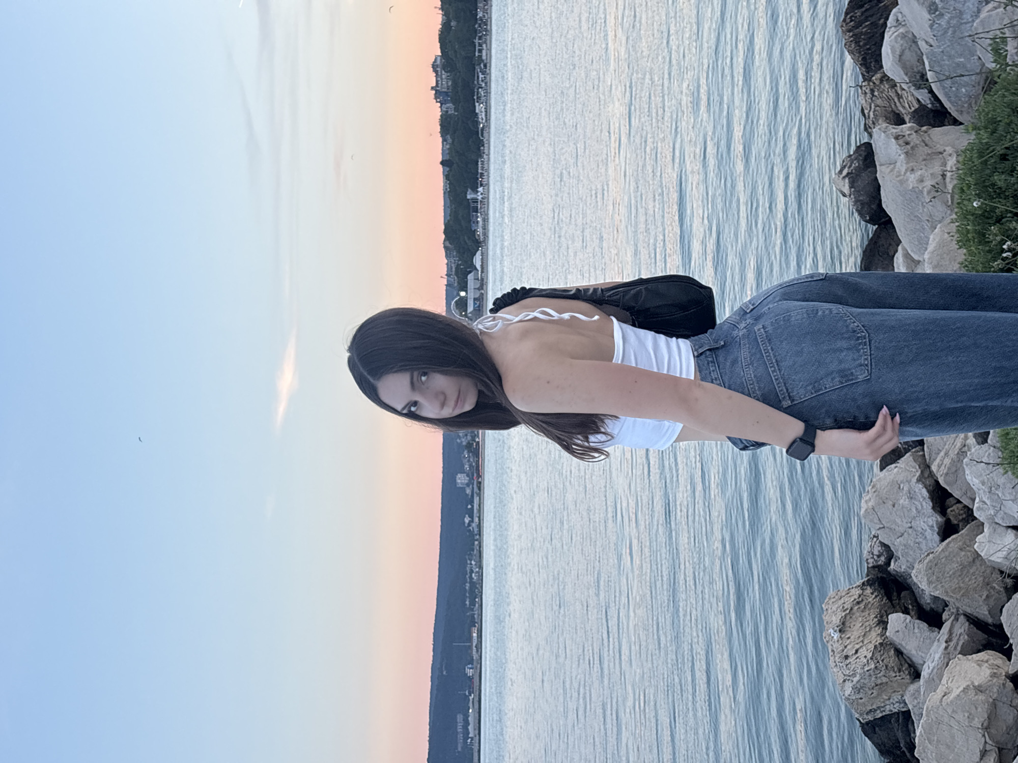 A young woman with long, dark hair stands on a rocky shoreline in Varna, Bulgaria, looking over her shoulder at the camera. She is wearing a white halter-neck crop top and high-waisted blue jeans. A black shoulder bag is slung over her right shoulder. She has a black smartwatch on her left wrist. The sea is calm with small waves, reflecting the pastel hues of the sunset sky. In the background, a distant coastline is visible with buildings and trees. The foreground consists of large, grey rocks and some sparse green vegetation. The overall scene suggests a peaceful evening by the Black Sea.