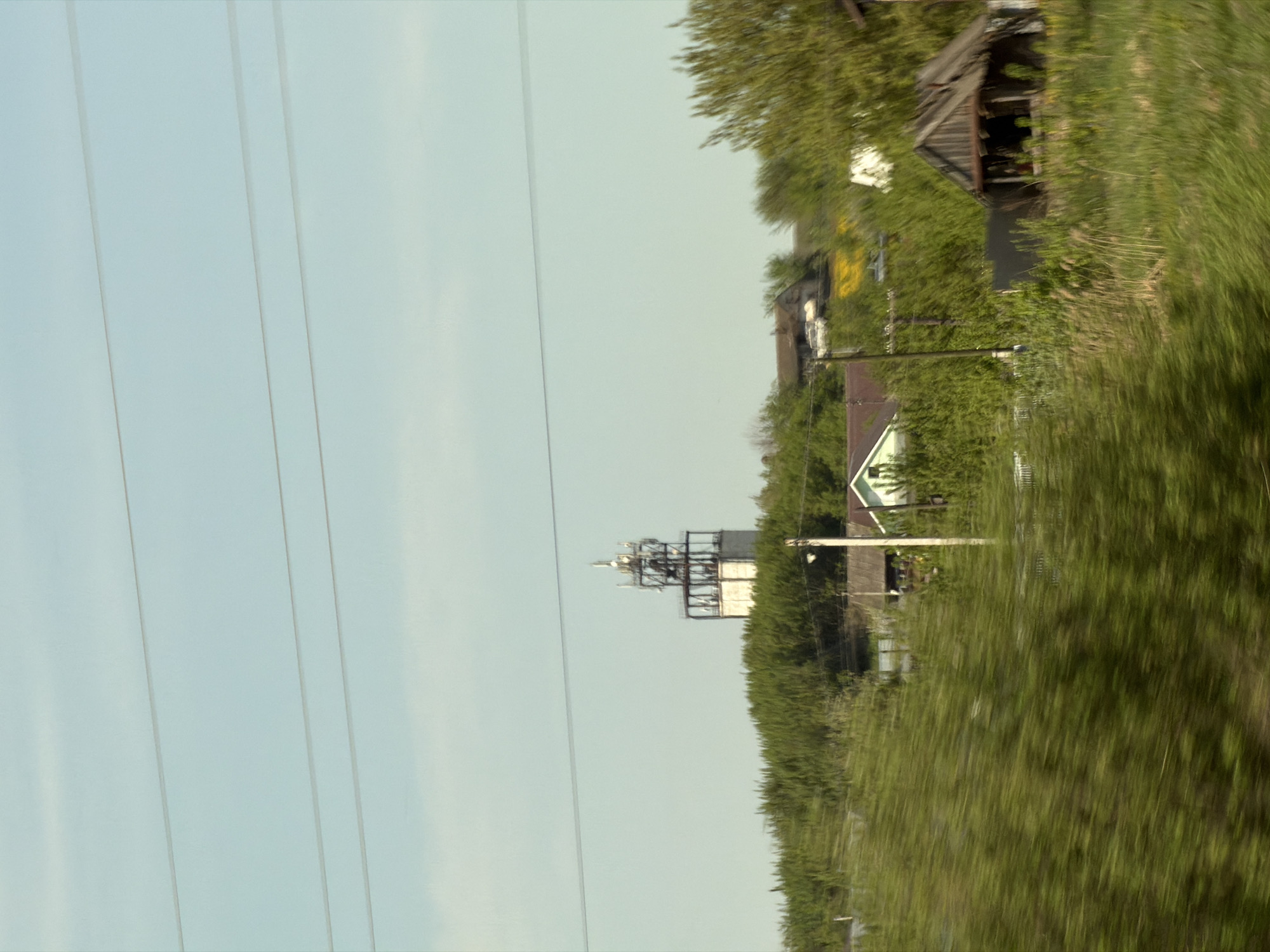 The image shows a blurred landscape near Совхозный, Russia. Power lines run across the left side of the frame. In the distance, a communication tower is visible. Vegetation and buildings are on the right, partially obscured by motion blur.