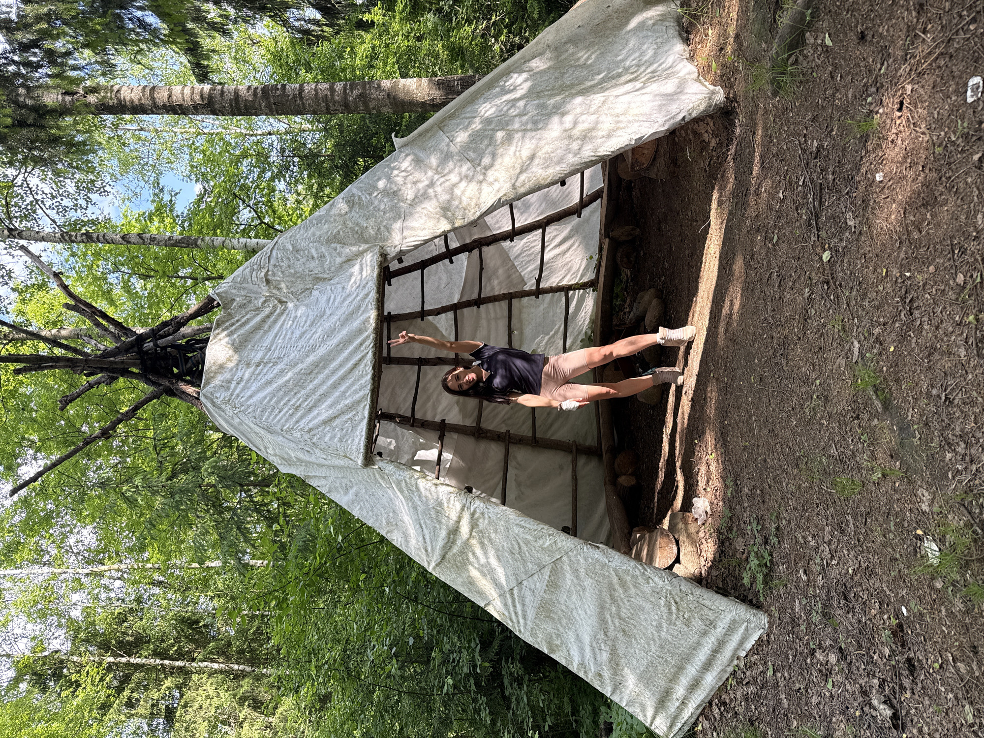 An outdoor, vertical photograph captures a person striking a pose within a rustic, temporary shelter in a sunlit forest. The setting is Sertolovo, Russia.
The primary subject is a woman, seen standing upright (though the image is rotated, presenting her horizontally) inside a simple structure. She wears a dark t-shirt, light brown or peach-colored shorts, white socks, and light-colored sneakers. Her dark hair is long, and she appears to be smiling or laughing. She stands on her left leg, with her right leg bent and raised slightly. Her left arm is bent at the elbow, hand raised, making a peace sign (V-shape with fingers). Her right arm is also raised.
The shelter is constructed from a white, possibly canvas or tarp-like material, draped over a framework of natural wooden poles and branches. The interior of the shelter shows more wooden supports and a dirt floor with some logs. The front of the shelter is open, allowing ample light to enter.
The background reveals a dense forest with tall trees, including birch trees with their distinctive white bark and other deciduous trees with lush green foliage, suggesting springtime or summer. The sky, visible between the tree canopies, is bright blue. The ground outside the shelter is a forest floor covered in dirt, dried leaves, and sparse green grass. Strong sunlight casts clear shadows, indicating a bright, sunny day.
No visible text is present in the image.