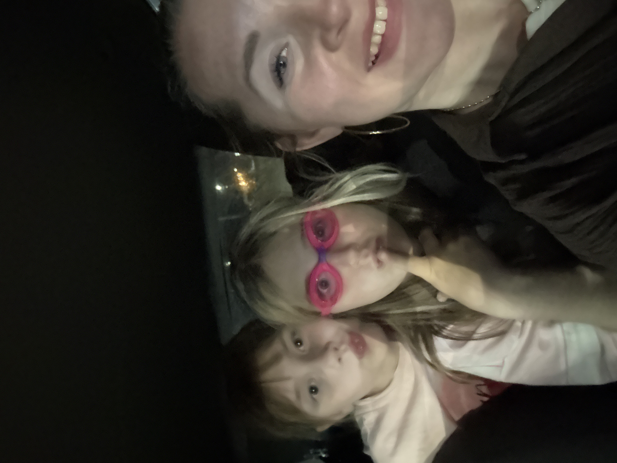In a dimly lit car at night, three females are captured in a selfie. The person in the foreground on the right, an adult female, smiles broadly at the camera, her face close to the lens. She has blonde hair, wears large hoop earrings, and a dark shirt. To her left, a young female is positioned in the middle, her face partially obscured by bright pink swimming goggles. She has blonde hair and her finger is near her mouth. On the far left, another young female is visible, her mouth pursed, and she is wearing a light pink shirt. The background is dark, suggesting the interior of a car, with some blurry lights visible through the rear window, hinting at an urban setting. The image does not contain any specific indicators of the city of Veliky Novgorod, Russia.