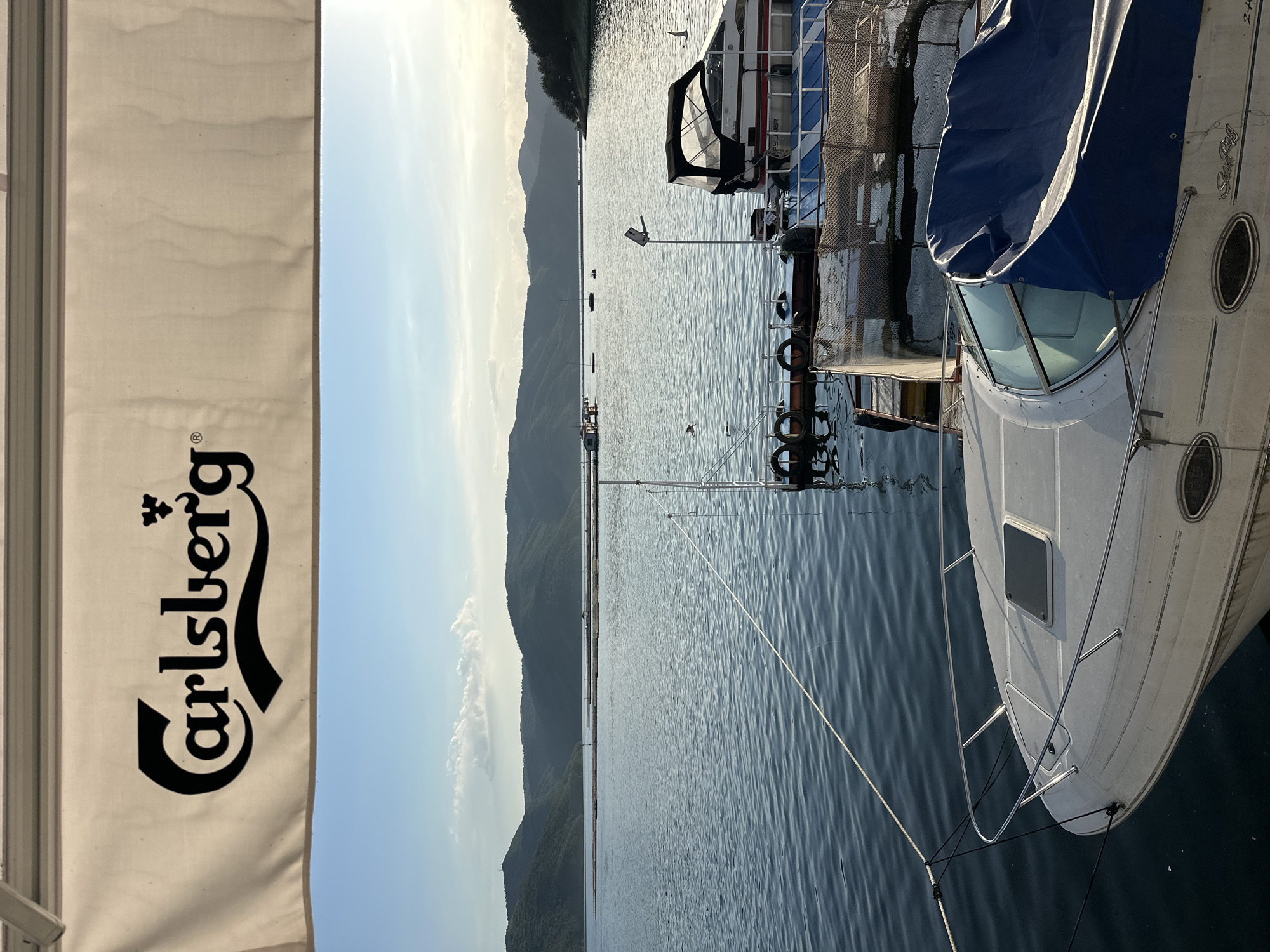 The image captures a tranquil outdoor scene in Brosh, Bulgaria, showcasing a large body of water framed by distant hills. The upper portion of the frame is occupied by a light-colored canvas awning, which prominently displays the black "Carlsberg" logo, including its stylized 'C' and a crown emblem above the 'g', along with a registered trademark symbol.
Below the awning, a wide expanse of calm water exhibits subtle ripples. In the immediate foreground, the white bow of a powerboat, partially covered by a dark blue tarp, is visible at the lower left. The name "Sea Ray" is faintly discernible on its hull. A white mooring rope extends from this boat towards a nearby pier structure on the right. This pier is constructed of wood or metal and features several black rubber tires used as fenders. Docked at the pier is a second white powerboat, equipped with a black soft-top canopy, oriented towards the right of the frame.
Further out on the water, multiple linear formations of low-lying structures, consistent with aquaculture pens or platforms, stretch across the mid-distance. Several smaller boats are anchored beyond these structures. The background is dominated by a continuous series of verdant, tree-covered hills or mountains, which gradually recede into the horizon. The sky is a clear, light blue, with a few scattered white cumulus clouds present, primarily above the hills. The scene is illuminated by natural sunlight, indicating either an early morning or late afternoon setting. No individuals are visible in the image.