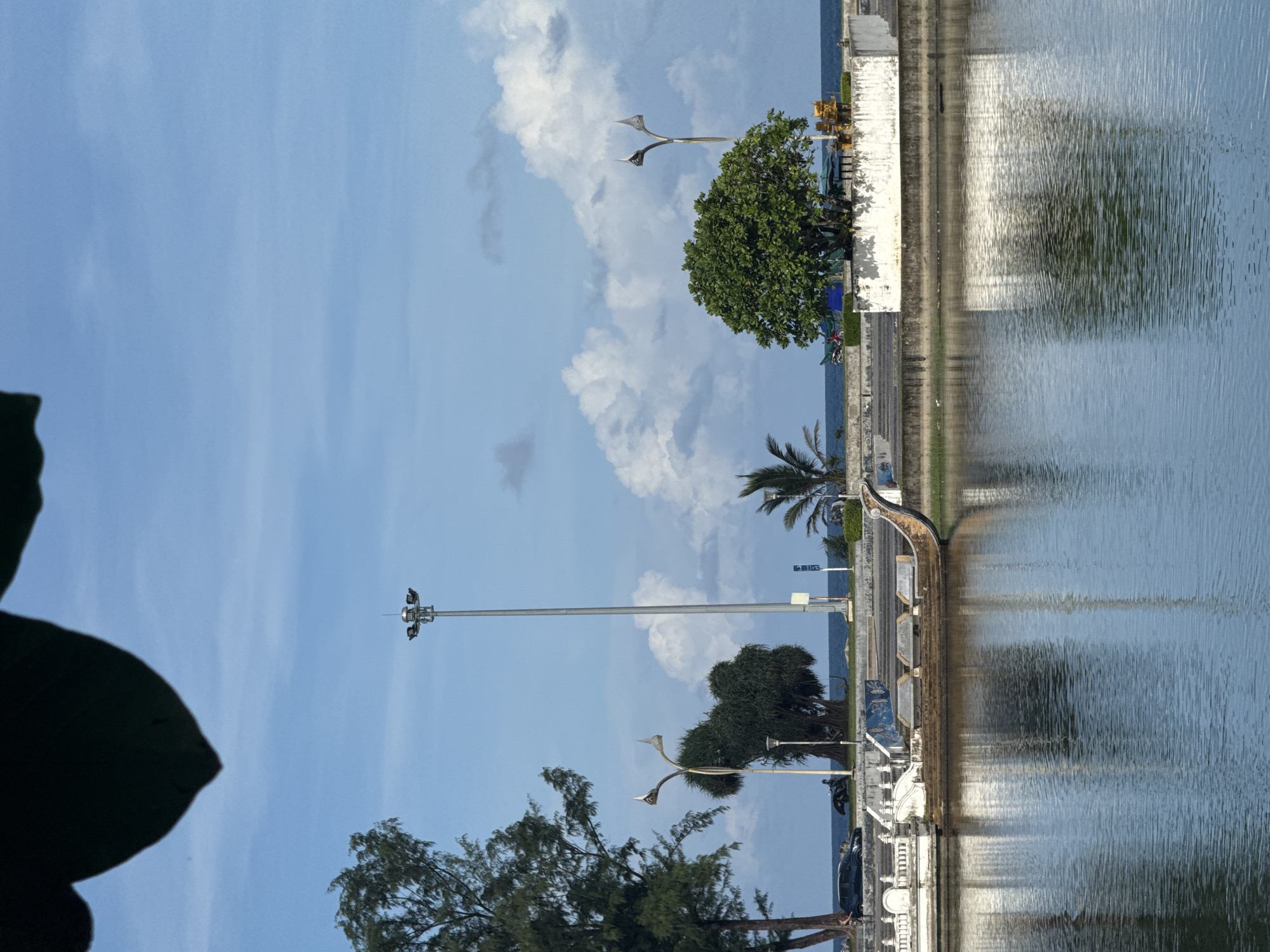 This is a full-frame, eye-level, medium shot of a waterfront scene in Karon, Thailand. In the foreground, a body of water with ripples reflects the sky and the scene behind it. A decorative, boat-like structure with ornate railings and benches sits on the edge of the water. Beyond this, a concrete embankment lines the water, with a paved road and sidewalk.
On the sidewalk, a tall, grey lamppost with multiple lights stands prominently. To its left, a dense green tree grows, its branches reaching towards the sky. Further left, another tree with a more sparse canopy is visible. In the distance, palm trees dot the landscape, and the blue ocean stretches to the horizon, meeting a sky filled with white, fluffy clouds. A few vehicles are visible on the road, including a dark-colored car and a motorcycle. The overall atmosphere is bright and somewhat serene, with a hint of tropical ambiance.