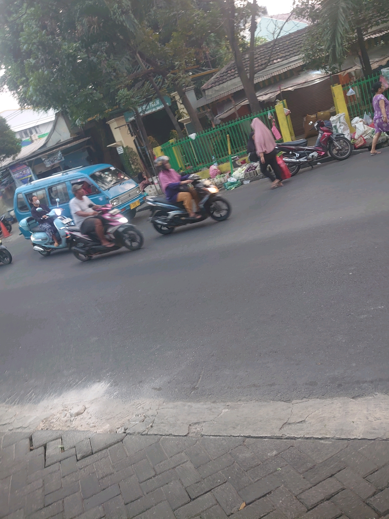 This is a medium-angle, outdoor shot of a street scene in South Jakarta, Indonesia, likely during the daytime. The perspective is slightly tilted, giving a dynamic feel to the image. The foreground is dominated by an asphalt road with a cracked concrete and brick sidewalk on the left. The road itself shows signs of wear and tear, with a lighter colored painted line partially visible.
In the mid-ground, several vehicles and individuals are captured in motion, indicating a busy urban environment. A light blue van is parked on the left side of the road. To the right of the van, a person is riding a light blue scooter, wearing a patterned outfit. Further to the right, a person in a pink hijab and dark clothing is walking alongside a dark motorcycle. Another person on a dark motorcycle, wearing a purple shirt, is riding in the same direction. Behind them, a red motorcycle is parked. On the far right, a person in a purple patterned outfit is walking away from the scene. Several green metal fences run along the background, separating the street from buildings and vegetation.
The background consists of buildings with tiled roofs, trees with dense foliage, and some market stalls or shops. The overall impression is one of everyday street life in a bustling South Jakarta neighborhood. The lighting suggests it is a sunny day, with some shadows present. The image is slightly blurred, likely due to motion or the camera's focus.