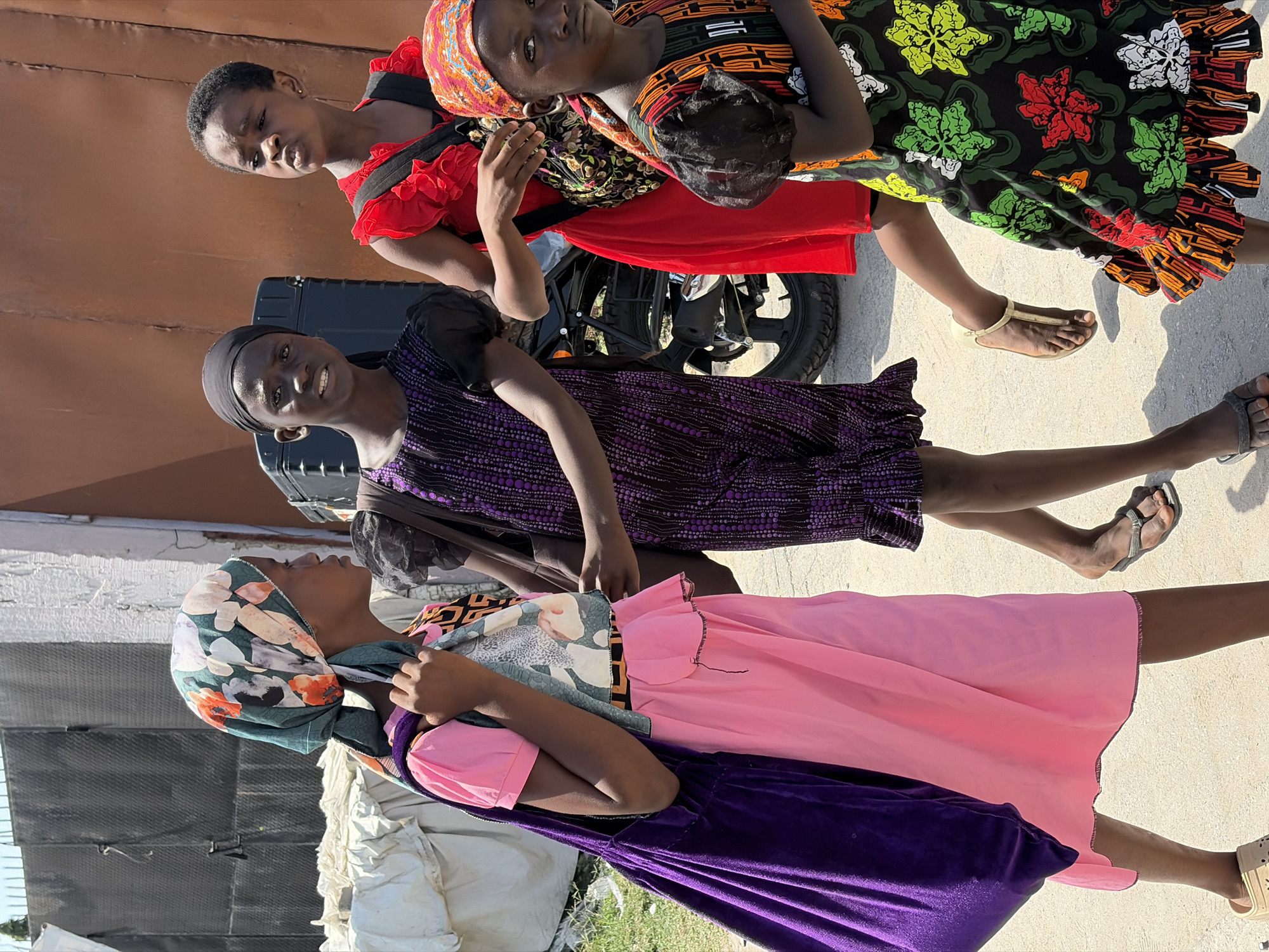 Three young women are standing outside in Igboefon, Nigeria. The woman in the front is wearing a pink dress with a purple velvet overskirt, and a floral headscarf. The woman in the middle is wearing a patterned purple dress and a black headband. The woman in the back is wearing a red dress and a patterned headscarf. A motorcycle is parked behind them, and there is a building in the background.