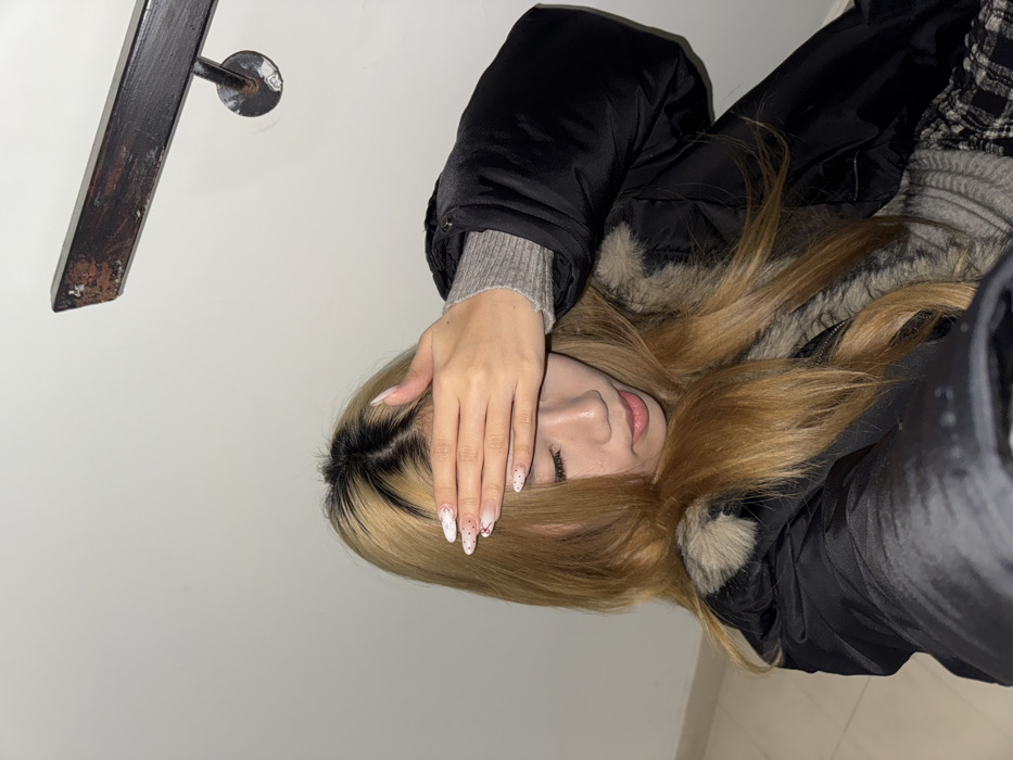 A close-up, selfie-style photograph depicts a young East Asian female indoors, positioned against a plain white wall. A dark, rectangular metal handrail, showing signs of rust or peeling paint, is mounted on the wall in the upper right quadrant, secured by a circular bracket. The subject's head is tilted downwards, and her right hand covers her forehead, obscuring her eyes. She has long, light brown hair with distinct dark roots. Her fingernails are long, pointed, and feature white tips with small red or white speckled patterns. She is wearing a black puffer jacket with a light brown/grey faux fur collar or trim, a grey ribbed knit sweater, and a garment with a black and white plaid pattern, possibly a skirt or pants. The lower left portion of the frame hints at a light-colored floor surface. No specific visual cues within the image identify the location as Yuzhong District, China.