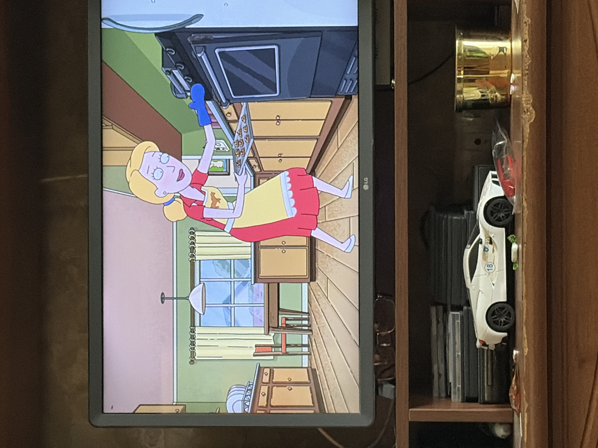 An animated woman with blonde hair tied in a ponytail is standing in a kitchen, placing a tray of cookies into an oven. She is wearing a red shirt, a yellow apron with a horse emblem, and a blue oven mitt on her right hand. The kitchen has light green walls, a wooden floor, and light brown cabinets. A dining table and chairs are visible in the background, along with a large window. The television screen is placed on a wooden shelf, below which are stacks of DVDs, a white toy sports car with the number 18 on it, and a gold cylindrical object.