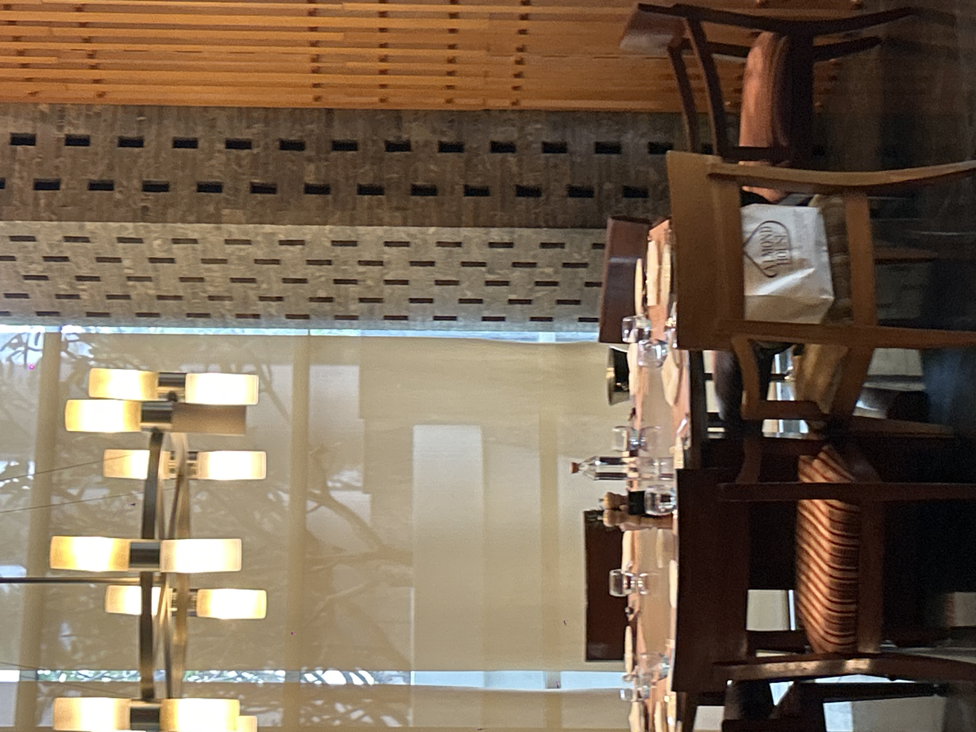 The image depicts an interior dining scene, likely a restaurant, with a focus on a round table set for a meal. The table is covered with placemats and has several place settings, including plates, glasses, and cutlery. A bottle of water and salt and pepper shakers are visible on the table. Surrounding the table are several chairs, one of which in the foreground has a striped cushion and a white paper bag with "ALMOND HOUSE" printed on it. In the background, there are large windows with sheer curtains revealing subtle tree silhouettes. A distinctive architectural feature includes a pillar constructed of stacked, rough-hewn stones with recessed dark rectangular openings. To the right of this pillar is a wall covered with vertical wooden slats. A modern chandelier with multiple cylindrical light fixtures hangs from the ceiling above the table. The overall ambiance suggests a contemporary dining establishment. The presence of the "ALMOND HOUSE" bag suggests a possible connection to a bakery or food establishment of that name, which is a known chain with outlets in Pune, India. Given the style of the restaurant interior, it is plausible this image was taken at an "Almond House" outlet or a restaurant associated with it in Pune, India.
