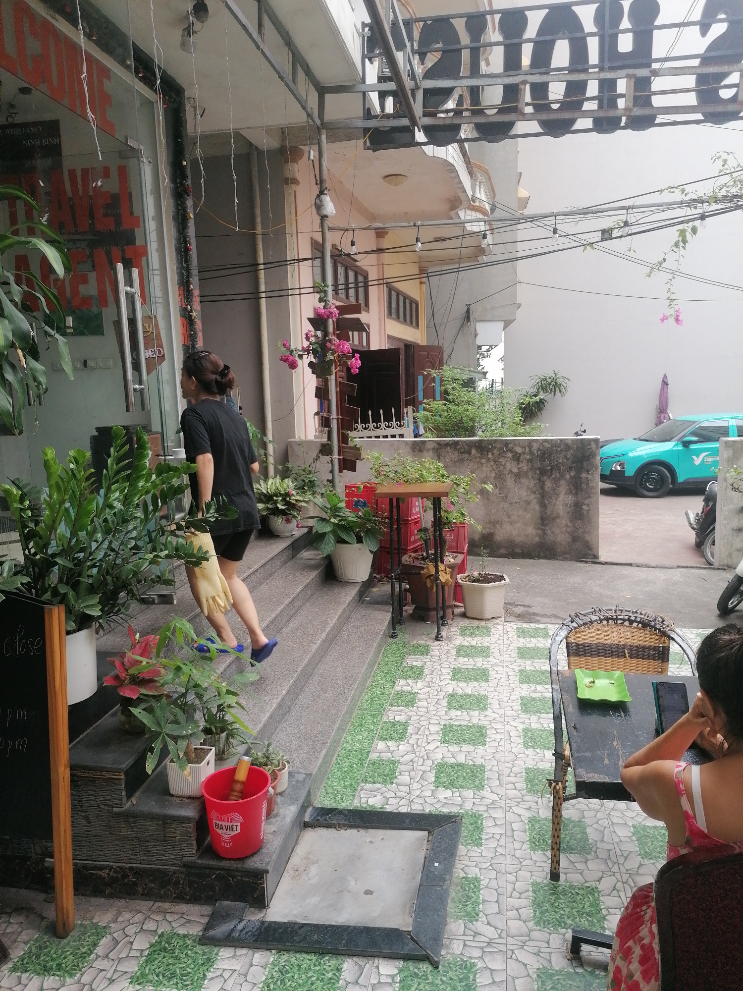 The image depicts an outdoor scene in Bai Chay Ward, Vietnam. A woman wearing a black t-shirt and shorts ascends a set of steps leading to a building with a "TRAVEL AGENT" sign on the glass door. She carries what appears to be a pair of yellow rubber gloves. Several potted plants decorate the steps.
To the right of the steps is a seating area with a tiled floor. A woman with short dark hair is seated at a table, appearing to be engaged with a phone or electronic device on the table. In the background, beyond a concrete wall, a light-blue car is parked. Electrical wires are visible overhead. The lighting suggests it is daytime. The surroundings are urban, as suggested by the building construction and the infrastructure.