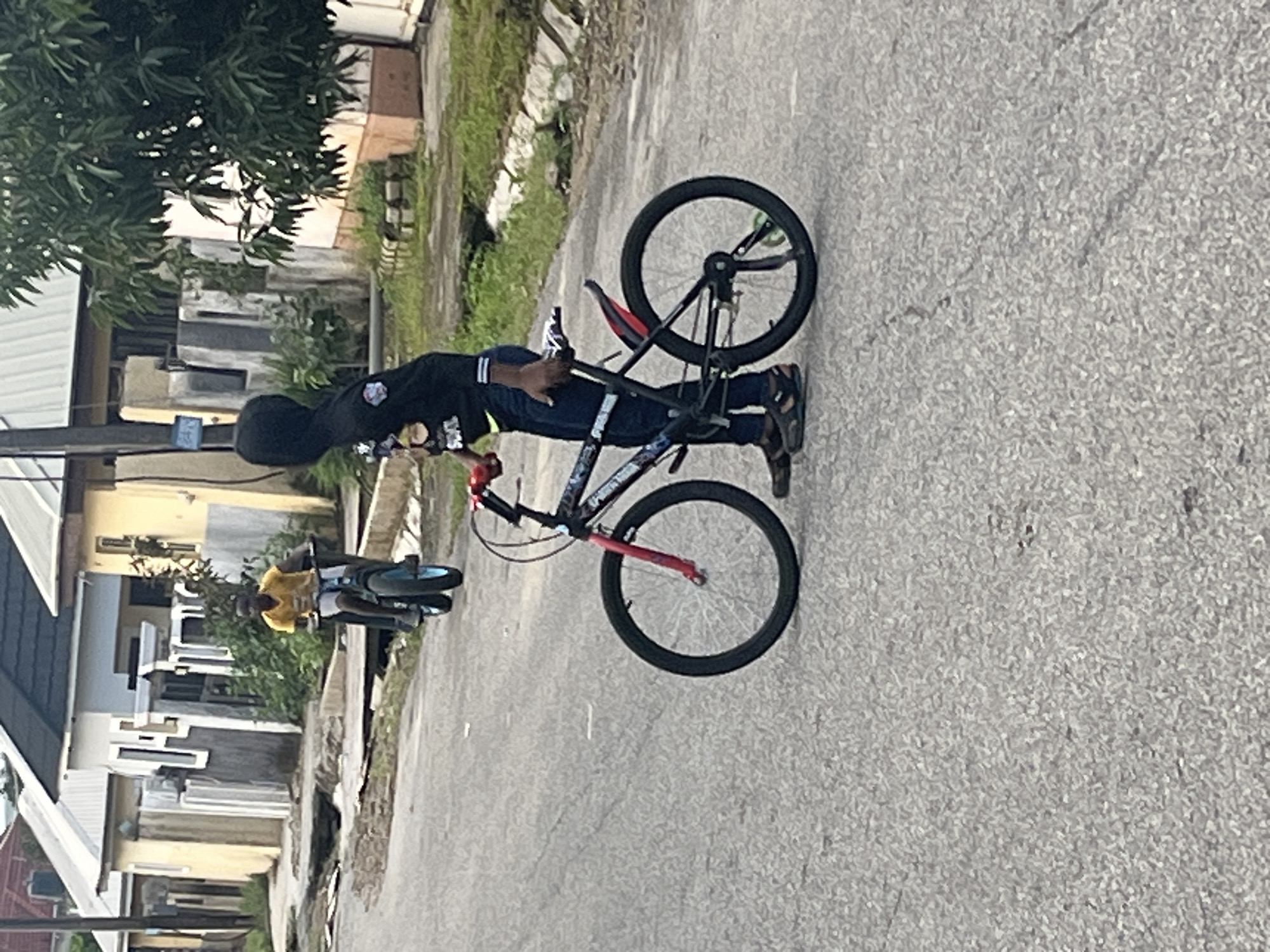 In Abuja, Nigeria, a young boy wearing a black hooded sweatshirt and blue jeans is standing next to a bicycle on an asphalt road. The bicycle has black tires with silver spokes, a black frame with red accents on the front forks and handlebars, and a black seat. The boy is holding the handlebars with his right hand. In the background, another boy on a bicycle is visible further down the road, wearing a yellow t-shirt and blue shorts. He is riding a bicycle with blue tires. The environment consists of residential buildings with cream and yellow facades, some with corrugated iron roofs, and overgrown vegetation. The road appears to be in moderate condition with some visible cracks.