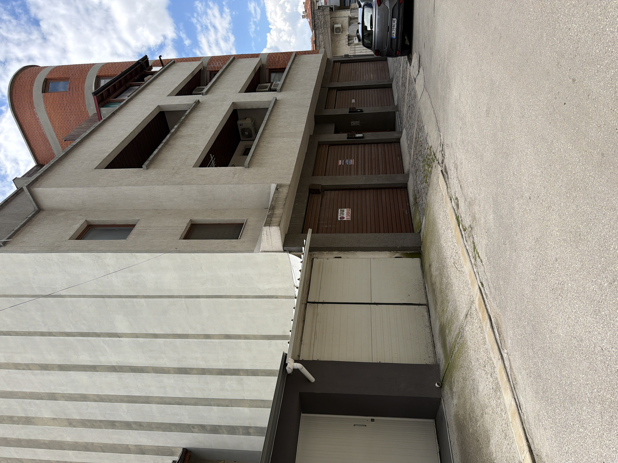 The image shows a row of garages and an apartment building in Pazardzhik, Bulgaria, under a cloudy sky. The building on the left has a grey facade with vertical ribbed detailing. To its right are two large white garage doors. A darker structure with a gutter system is attached to the left of these garages. The main apartment building is a multi-story structure with a beige facade, featuring balconies with wooden undersides. The rightmost section of the building is a curved structure faced with red brick and concrete bands, with a small window visible. Below the balconies are multiple brown roll-up garage doors. A dark gray car with license plate "PA 3946 ME" is parked on the right side of the image, facing away from the viewer. The ground in front of the garages and leading to the street is paved with concrete and cobblestones. There are two signs on the brown garage doors indicating "GARAGE" and "PARKING".