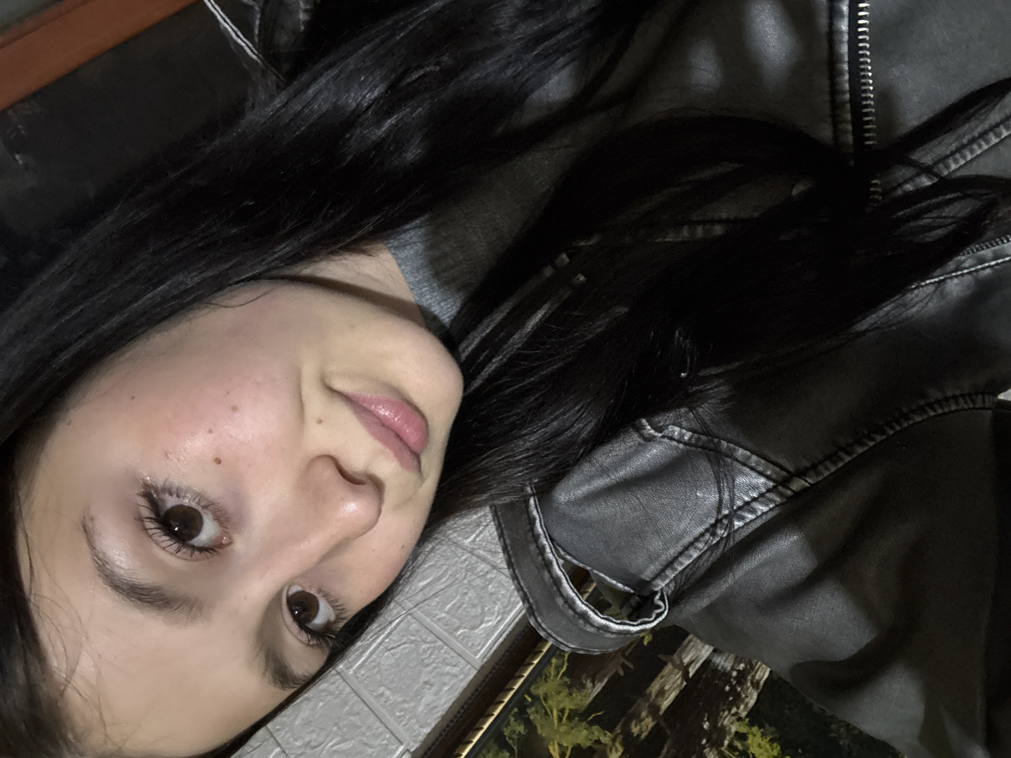 A young woman with dark hair is posing for a selfie. She is wearing a black leather jacket over a gray top. The background appears to be an outdoor space in the evening, with a brick wall visible in the bottom left corner. Vegetation can be seen in the lower right corner, hinting at a possible garden or park area. It is not possible to ascertain if the picture was taken in Фоки, Russia.