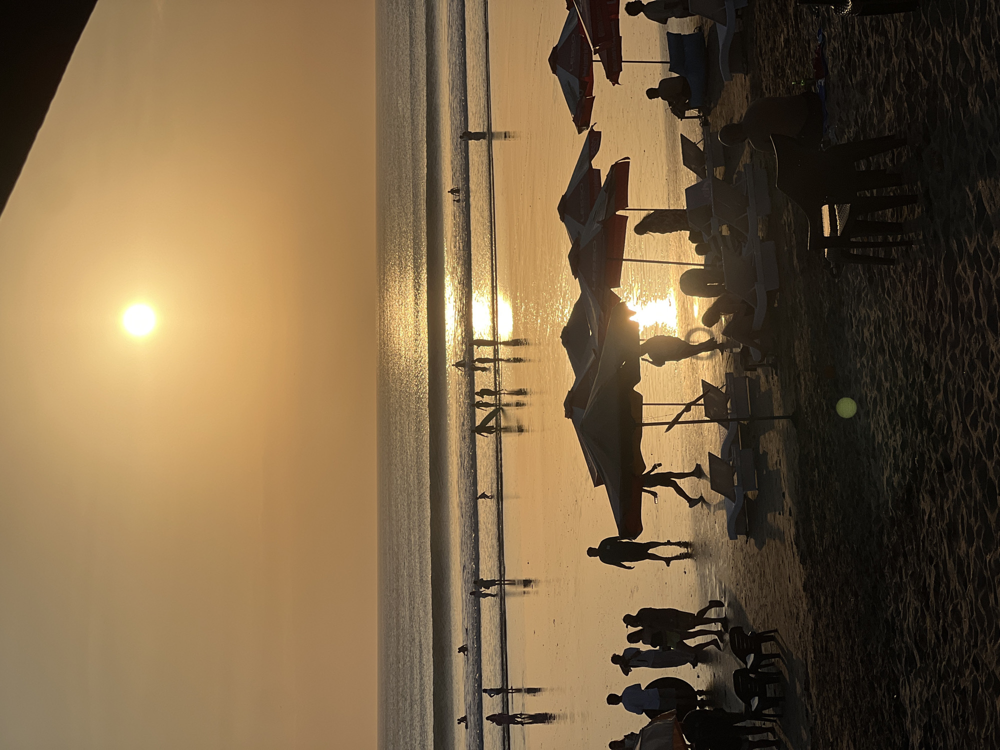 The image captures a vibrant sunset scene on Kuta Beach, Bali, Indonesia, with the sun low on the horizon, casting a warm, golden glow across the sky and water. Silhouetted figures dot the beach and the shallow water, engaged in various activities. Several people are standing on the wet sand, some walking, while others appear to be wading or playing in the gentle waves. A group of individuals is visible in the ocean, some with surfboards, suggesting they are surfing or about to surf. In the foreground, beach umbrellas and lounge chairs are set up on the sand, some occupied by people relaxing. The sand is textured with ripples from the wind. The ocean exhibits rolling waves, reflecting the intense sunlight, creating a shimmering effect on its surface. The overall atmosphere is one of leisure and natural beauty, typical of a popular tourist destination at dusk.