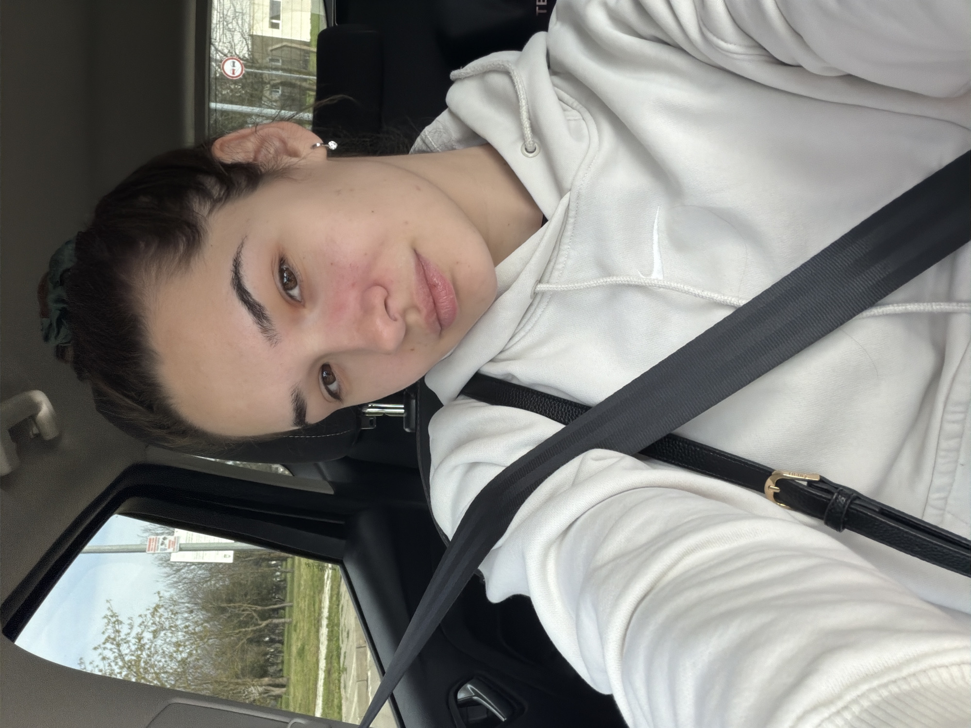 A medium shot, slightly angled, shows a young adult female in the driver's seat of a car. She has brown hair, brown eyes, and is wearing a white Nike hoodie. A black seatbelt crosses her chest. Outside the car window, in daylight, is a street scene with bare trees and green grass. A street sign is partially visible. The location is Kavarna, Bulgaria.