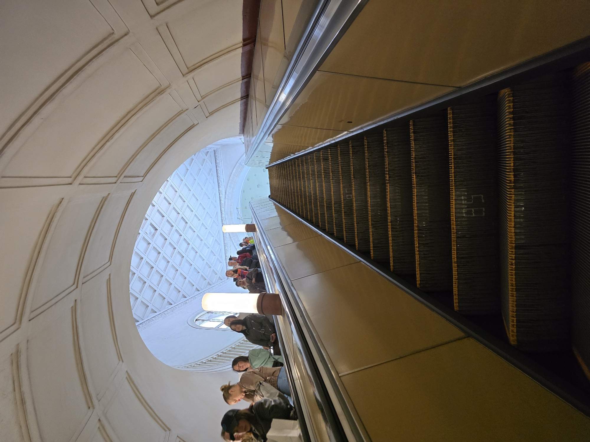 This photograph, taken in Moscow, Russia, depicts an escalator ascending through a subway station. The escalator's steps are black with yellow stripes. People are riding the escalator upwards. The architectural design features curved white walls with gridded patterns and oval windows. Elongated, cylindrical lights are suspended above the escalator.