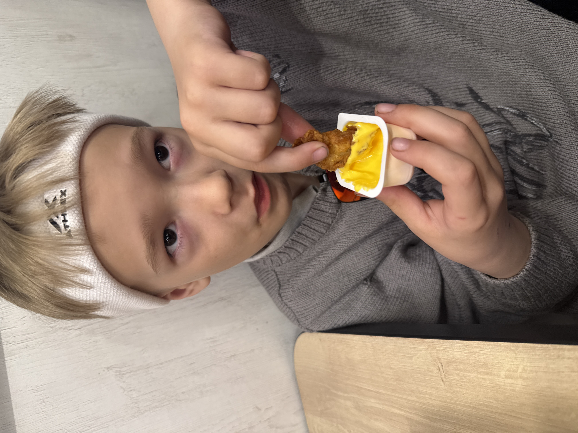 A child wearing a white headband with black designs is shown from the chest up, holding a chicken nugget in their right hand and dipping it into a container of yellow sauce held in their left hand. They are wearing a gray sweater and are seated at a table. The background includes a light-colored wooden surface. Based on prior knowledge and the analysis of visual elements, the location is possibly in the city of Dmitrov, Russia, but this cannot be definitively confirmed from the image alone.