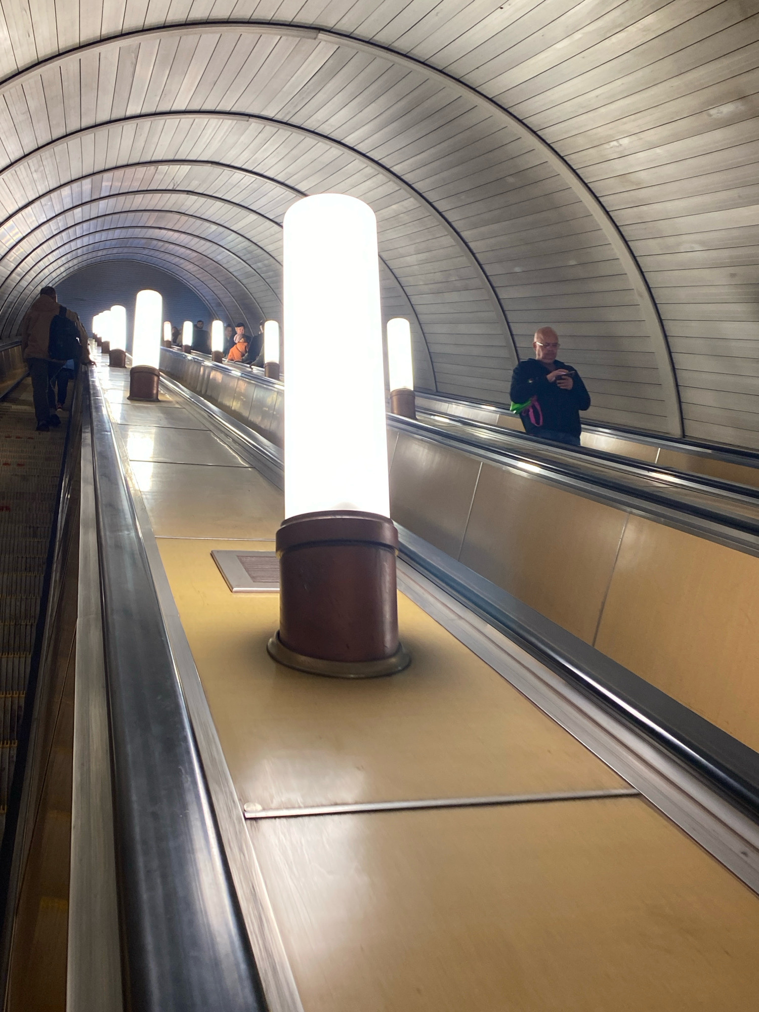 An escalator ascends through a curved tunnel lined with horizontal wooden panels and illuminated by vertical cylindrical lights. Several people are on the escalator. In the foreground on the left, a person wearing a brown jacket and a backpack is ascending. To the right, a bald man wearing glasses and a dark jacket is holding a phone. Other figures are visible further up the escalator. The environment appears to be a Moscow Metro station, likely one of the deeper stations with longer escalators.