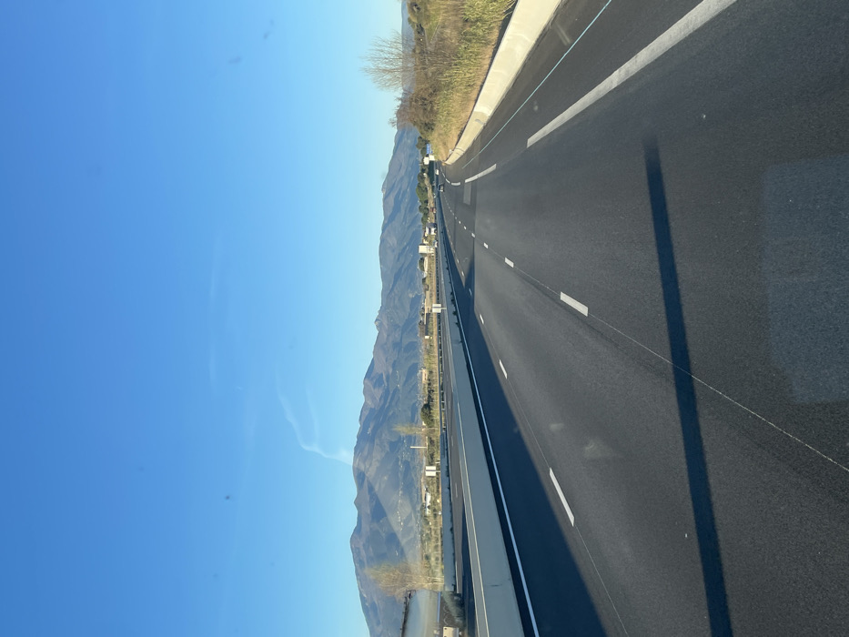 The image presents a forensic view of a multi-lane highway located within or near Le Boulou, France. The scene is captured under a clear, bright blue sky, with long shadows cast across the dark asphalt, suggesting either early morning or late afternoon.
The highway features at least two lanes of travel in the depicted direction, marked by white dashed lines, and is separated from opposing traffic by a concrete median barrier. A solid white line delineates the right shoulder, which is bordered by a concrete barrier and wild, dry vegetation including shrubs and grasses. Small white signposts are visible at intervals along the median barrier.
In the mid-ground to the left, a light-colored building or wall section is partially visible, along with several sparsely leaved deciduous trees. In the distance, a prominent range of mountains dominates the horizon. Several vehicles are observed traveling on the highway in both directions, appearing in the mid-ground and background. A faint, wispy contrail is present in the upper portion of the sky. No individuals are visible.